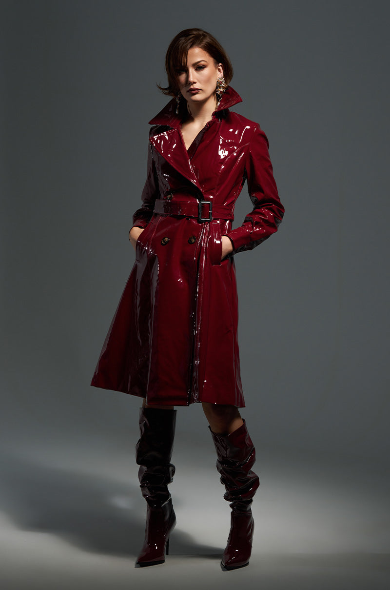 A person dressed in a MITICA PATENT TRENCH COAT, crafted from shiny burgundy faux patent leather, is paired with matching knee-high boots and stands against a plain dark backdrop. The coat displays double-breasted button closures and includes a belt, while they pose confidently with hands in pockets.