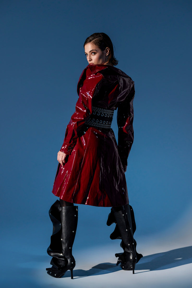 A woman stands against a blue background wearing the shiny red MITICA PATENT TRENCH COAT and black ruffled high-heeled boots, looking over her shoulder with a confident expression.
