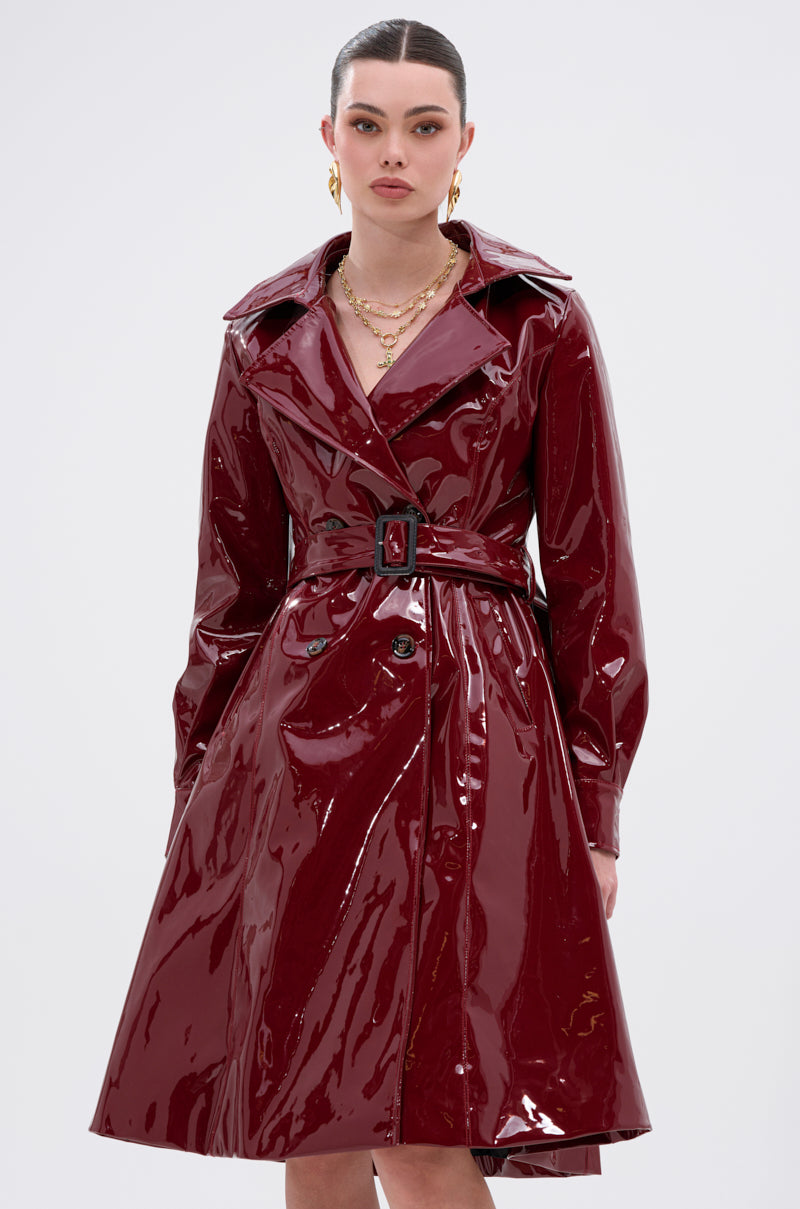 A woman models the MITICA PATENT TRENCH COAT—shiny dark red faux patent leather with double-breasted buttons—standing against a plain light background, accessorized with gold jewelry and looking directly at the camera.