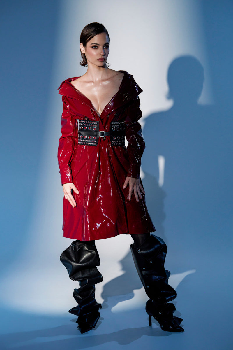 Model wears the MITICA PATENT TRENCH COAT—shiny red faux leather with studs and a belt—styled with oversized, slouchy black thigh-high boots. Blue-toned lighting casts dramatic shadows on the wall behind.