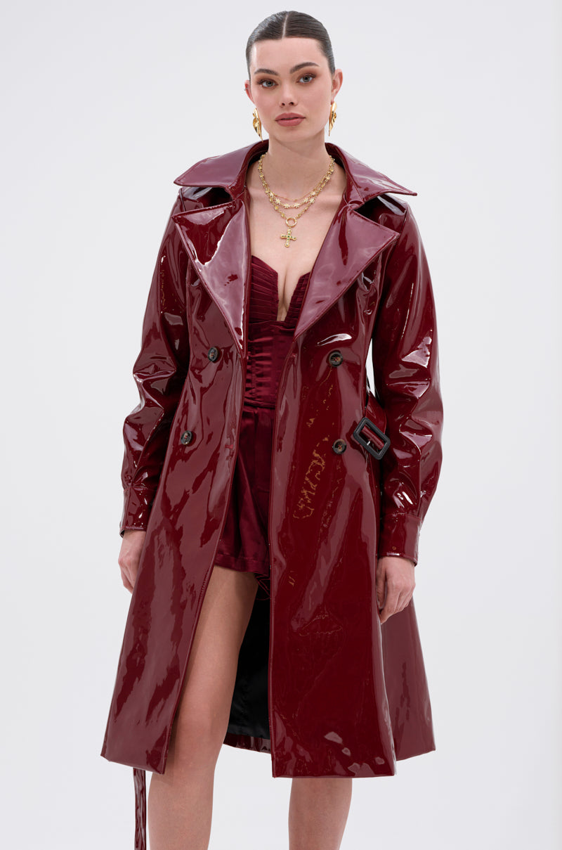 A woman models the MITICA PATENT TRENCH COAT in shiny burgundy faux patent leather with tortoise shell buttons, styled over a matching dress and gold jewelry, standing against a white background.
