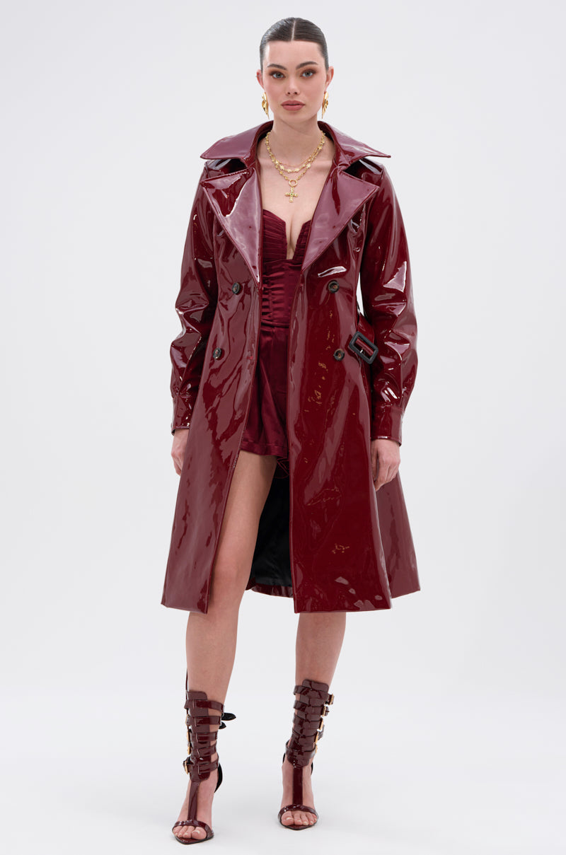 A woman poses against a plain background in the MITICA PATENT TRENCH COAT, a shiny burgundy faux patent leather piece with double-breasted buttons, styled over a matching ruched mini dress, strappy boots, and layered gold necklaces.