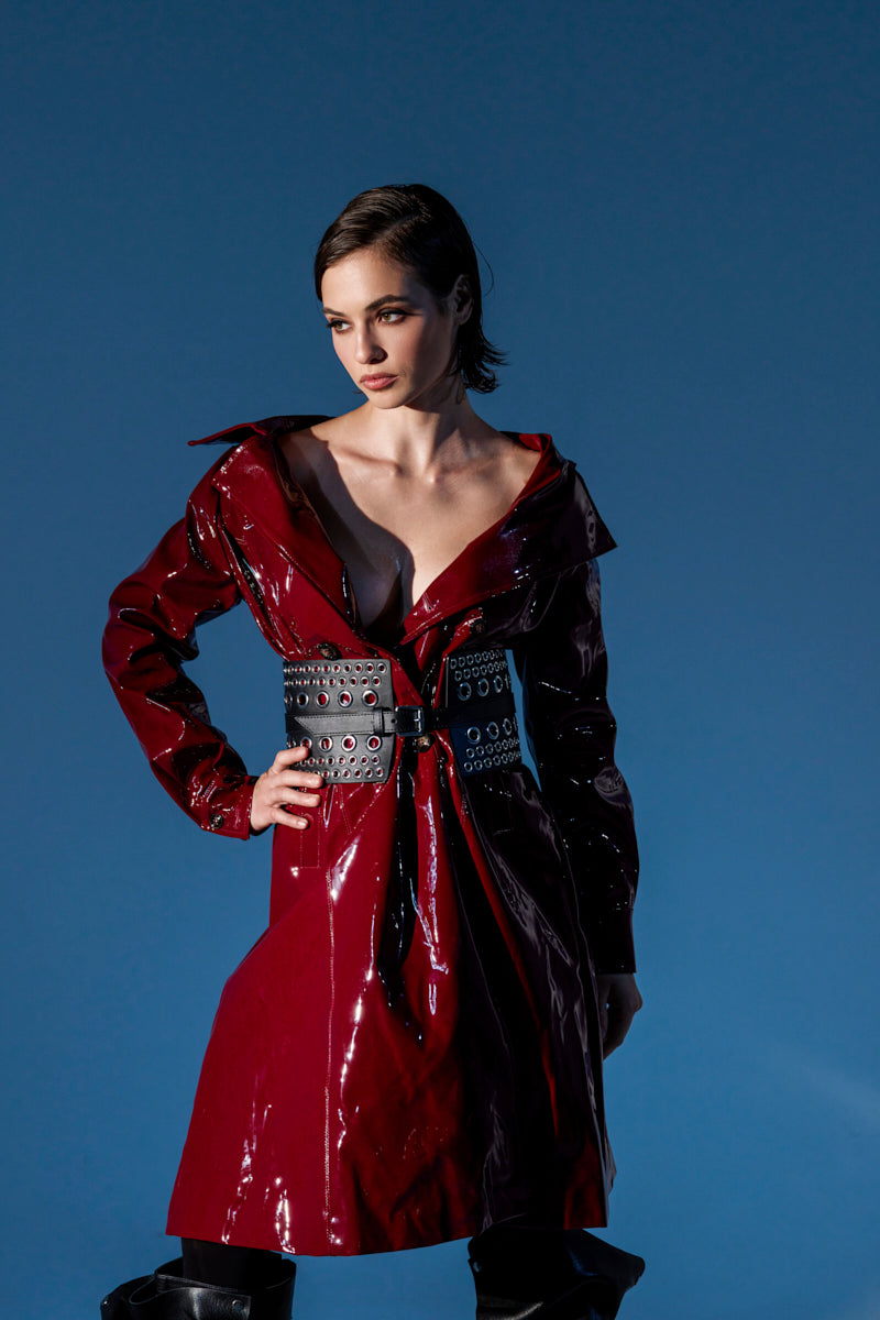 Confident woman in a MITICA PATENT TRENCH COAT with a studded black belt stands against a plain blue background, gazing to the side.