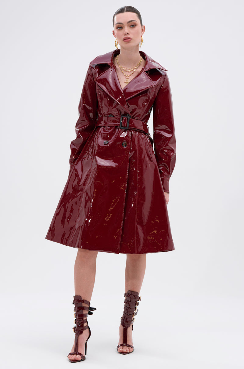 A woman models the MITICA PATENT TRENCH COAT in glossy dark red with a belted waist, styled with strappy heels, gold jewelry, and hoop earrings against a light background.
