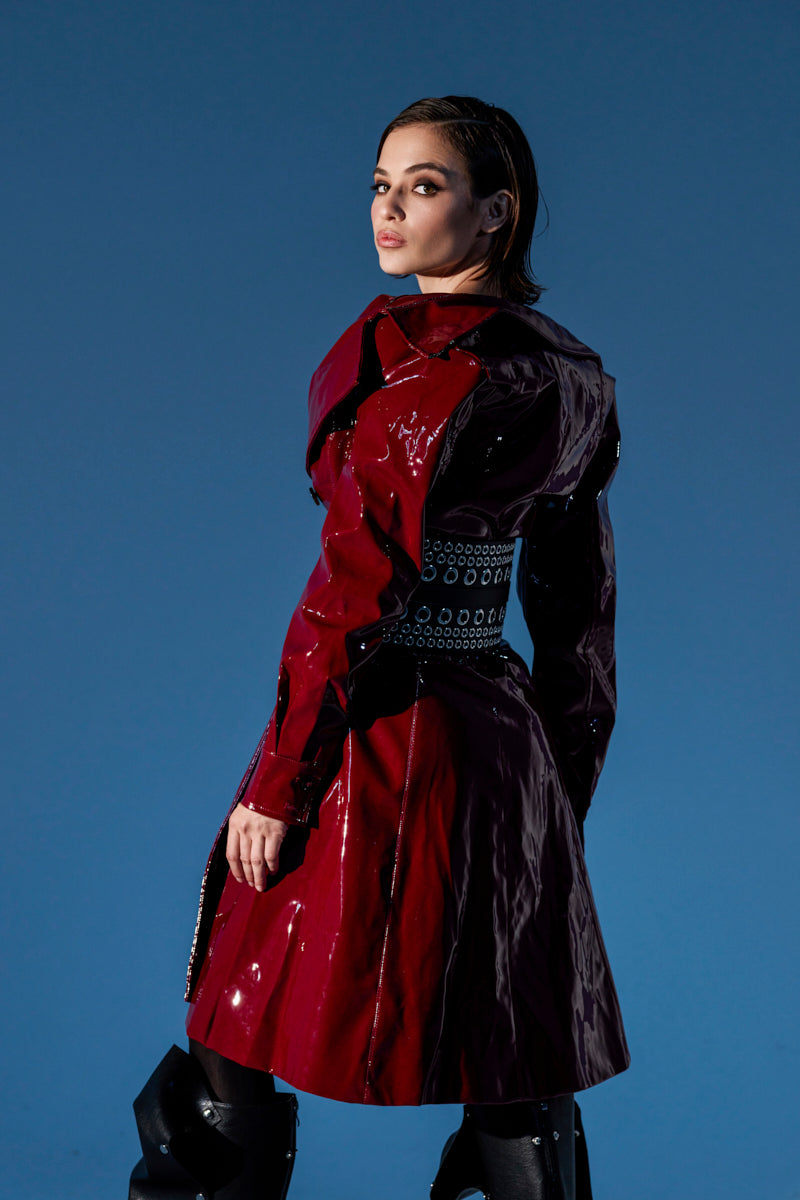 A woman stands confidently against a blue background, looking over her shoulder while wearing the MITICA PATENT TRENCH COAT in shiny red faux leather with a studded belt detail.