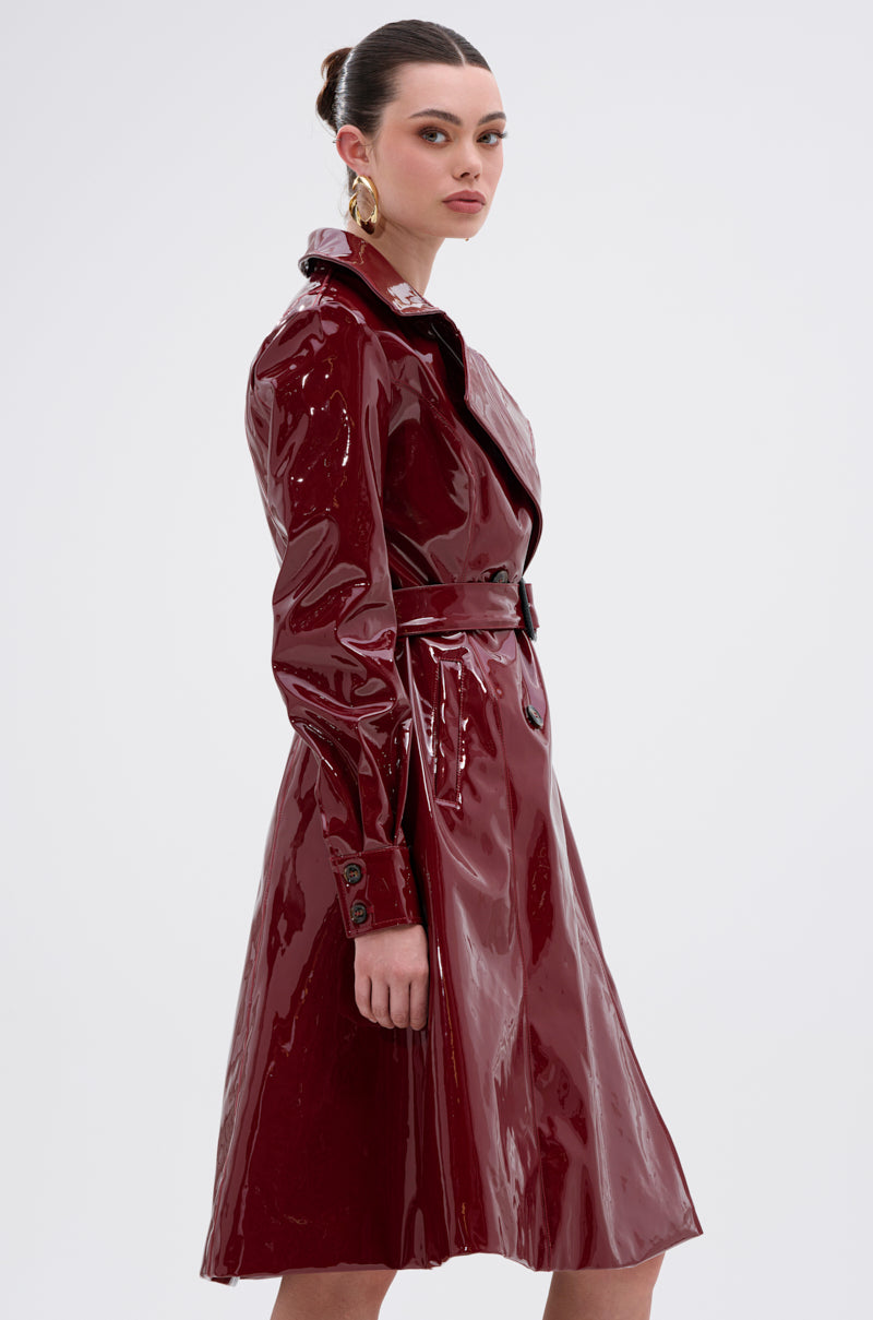 A woman, her hair in a neat bun, poses sideways against a plain background in the MITICA PATENT TRENCH COAT—crafted from glossy faux patent leather with double-breasted buttons—accented by gold hoop earrings as she glances over her shoulder.