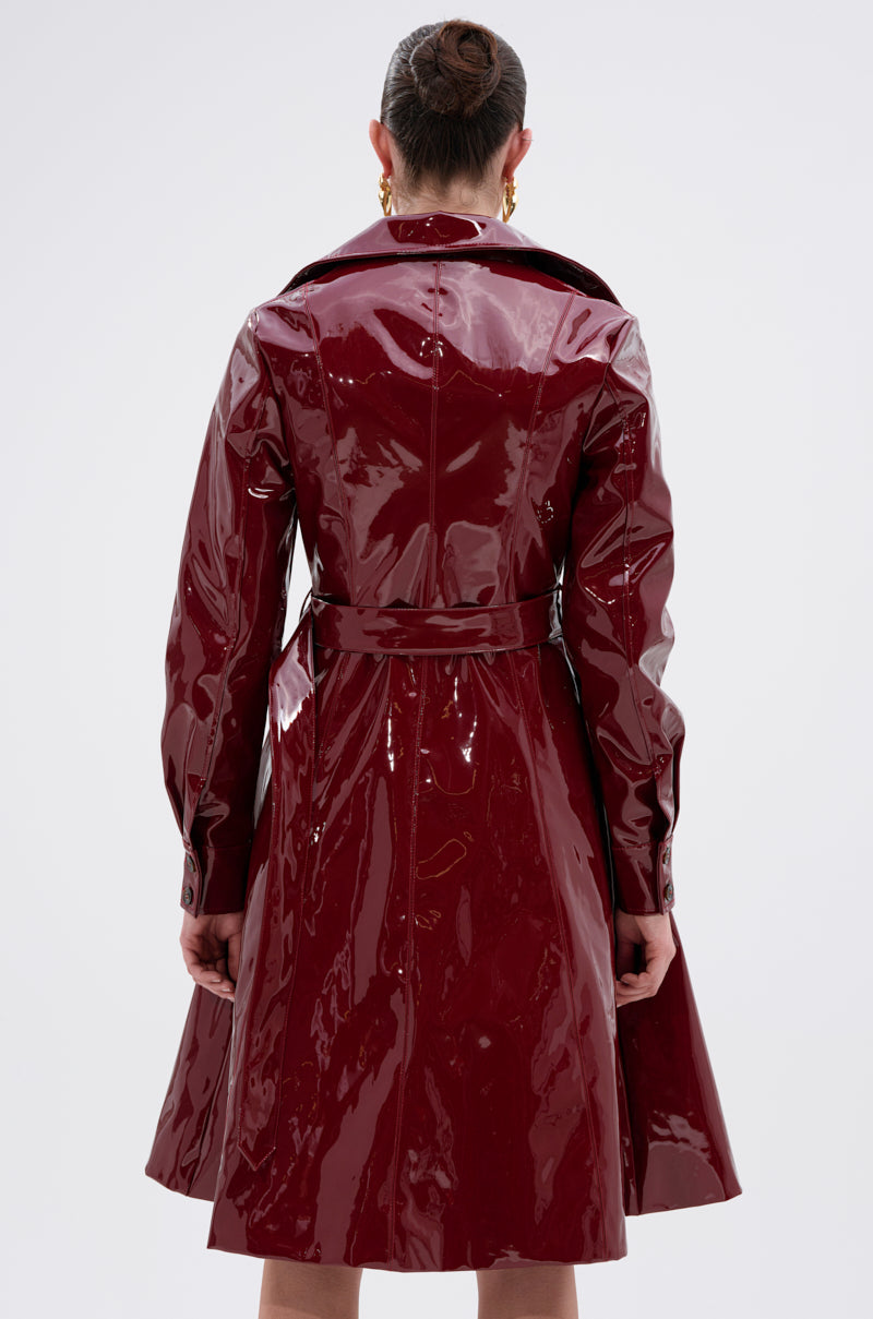 A person faces away, wearing the MITICA PATENT TRENCH COAT in shiny dark red faux patent leather with a belt and wide collar. Their hair is in a neat bun, gold hoop earrings shine, set against a plain light gray background.