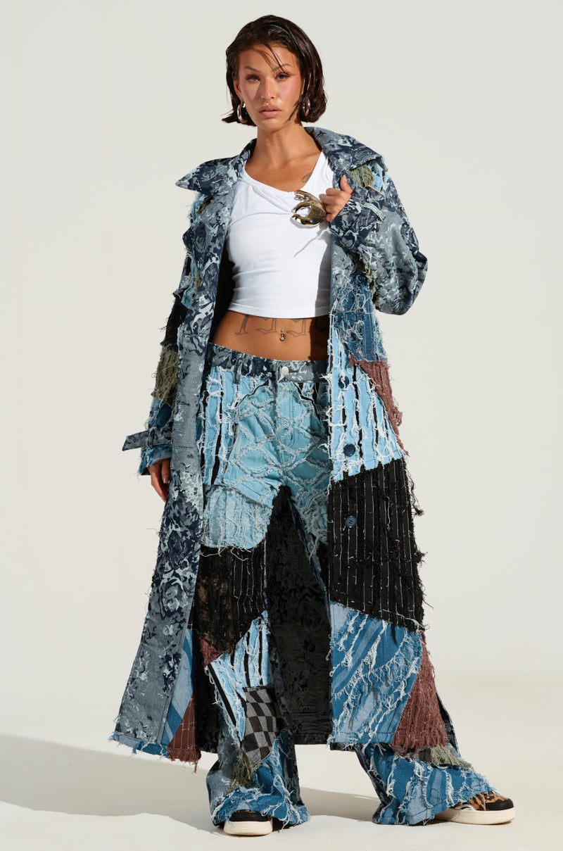 The individual is dressed in the RONNY PATCHWORK TRENCH, styled over a white top and matching pants. They are holding sunglasses and exuding confidence against a neutral backdrop.