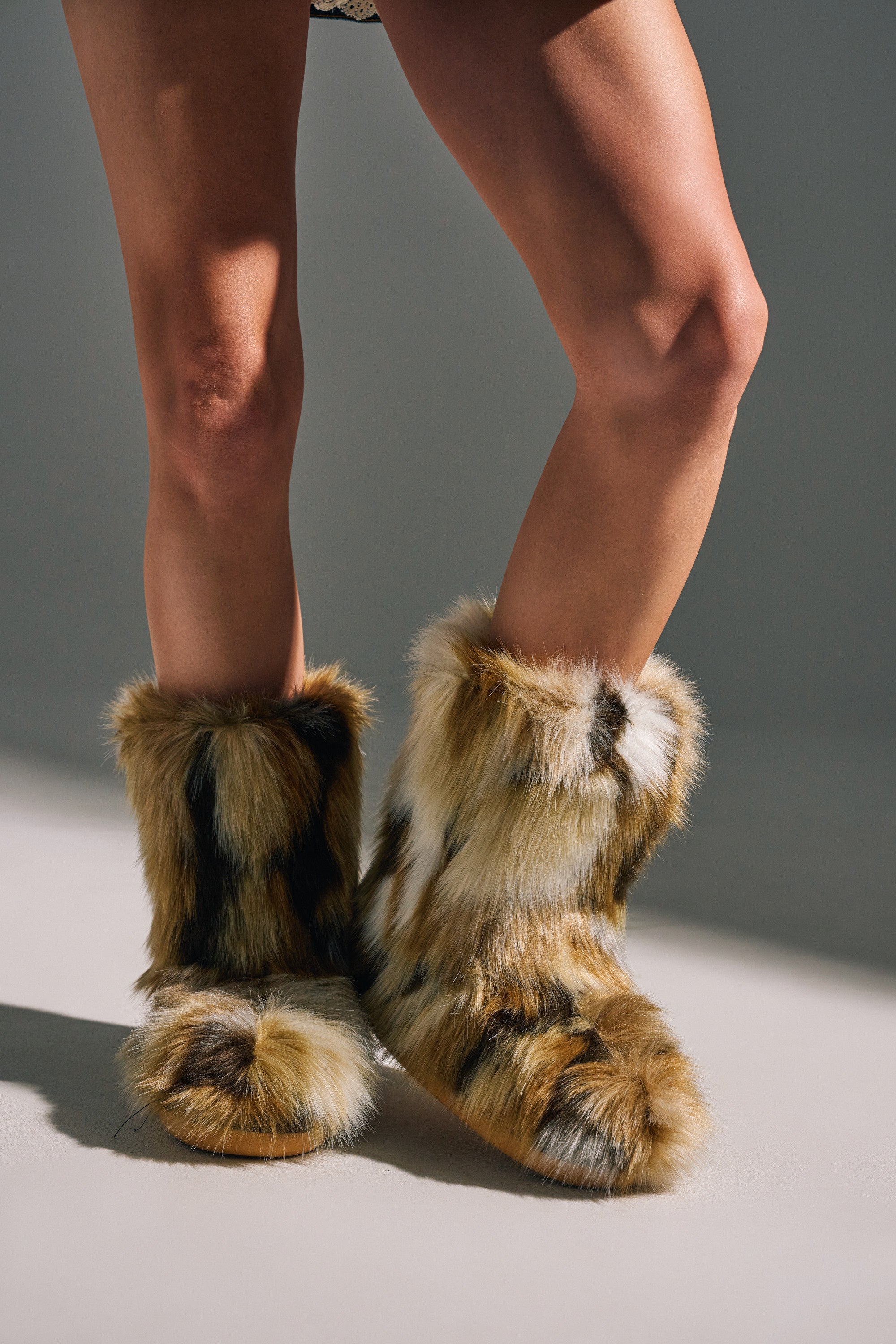 A person stands on a light surface, their legs and feet visible in AZALEA WANG NADIANNA BROWN FUR FLAT BOOTIE—tall, flat booties featuring brown, tan, and black spotted faux fur.
