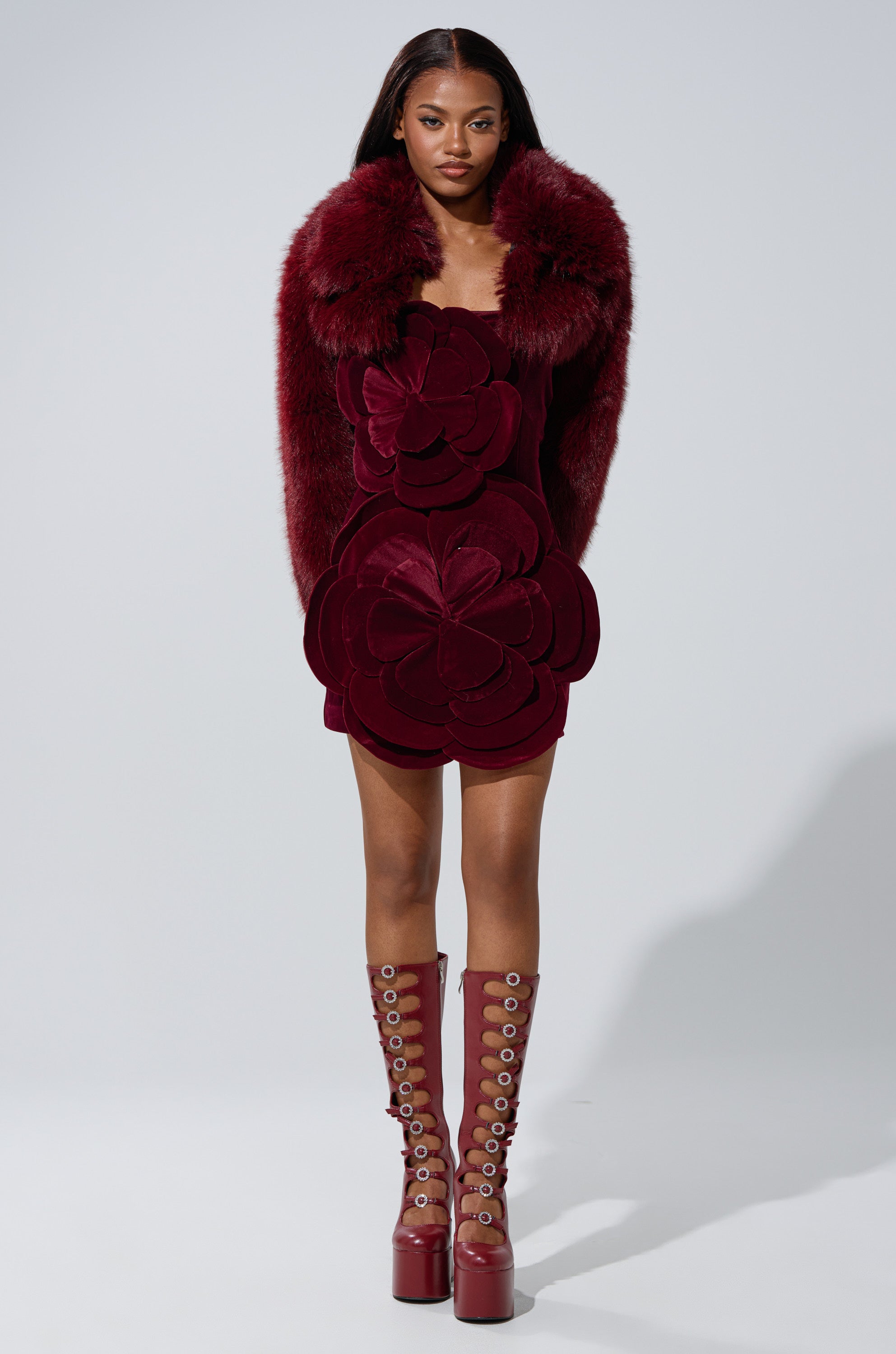 A woman poses in a deep red mini dress with large flower appliqués, the LEILA ULTRA CROP FAUX FUR IN BURGUNDY by AKIRA Label, and tall platform lace-up boots with circular cutouts. The background is plain and light gray.
