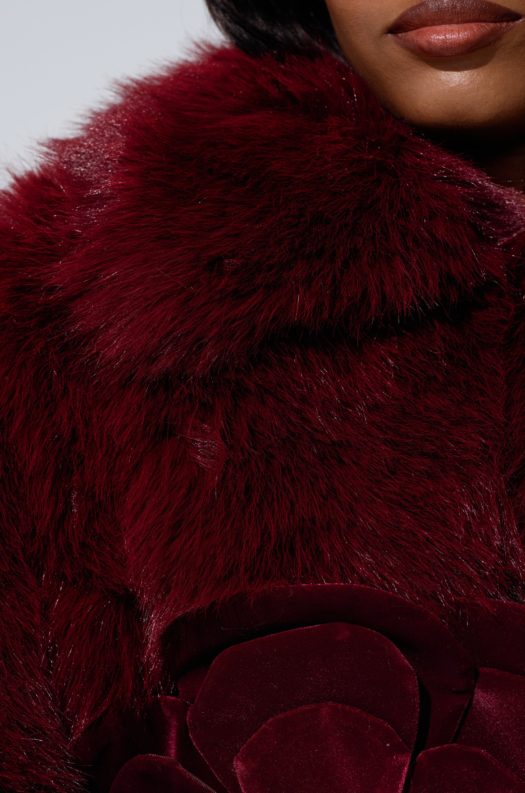 A close-up shows a person in the LEILA ULTRA CROP FAUX FUR IN BURGUNDY jacket, featuring large floral velvet detail; only the lower face and coat are visible.