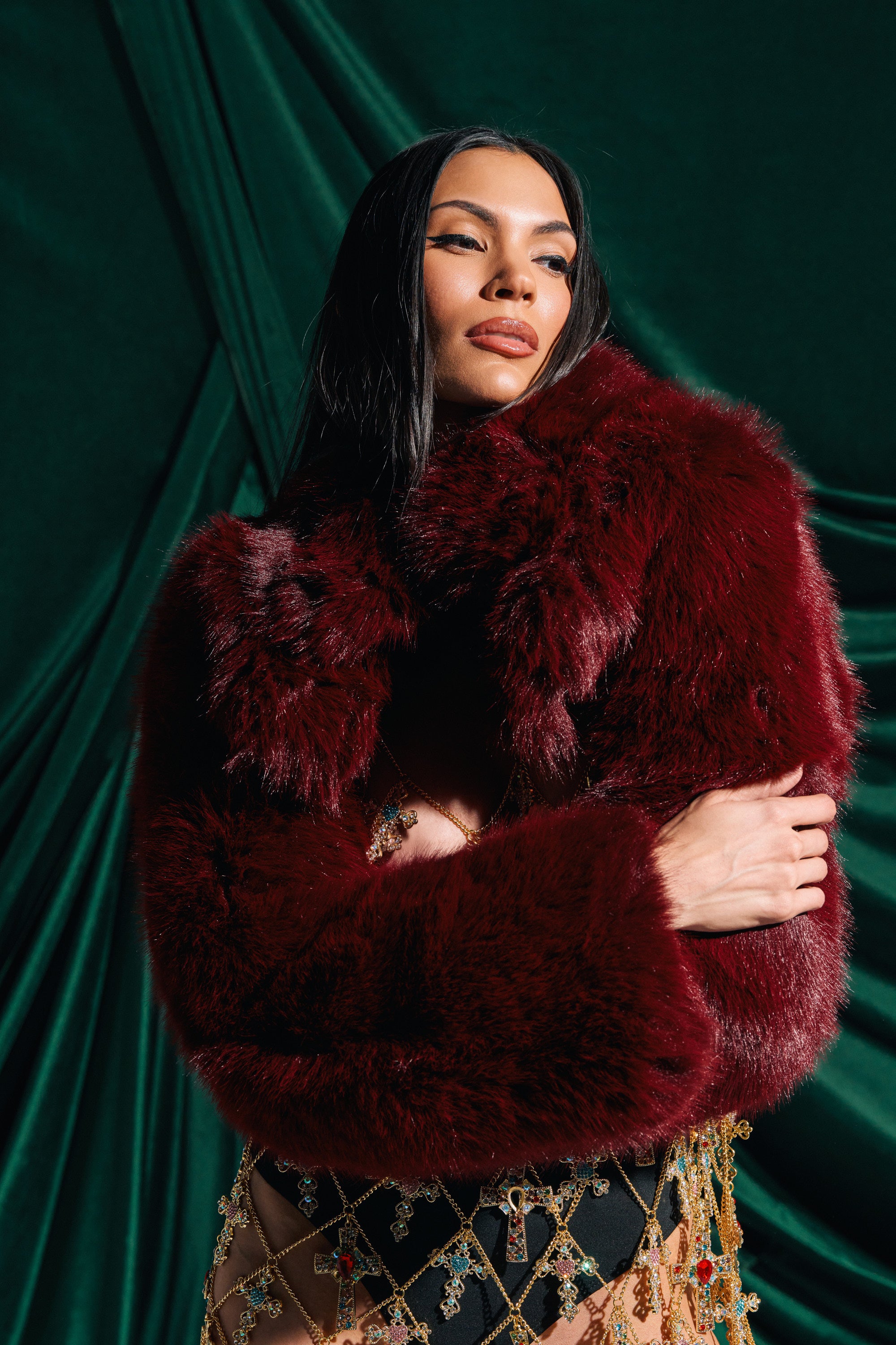 A woman with long dark hair poses confidently in the LEILA ULTRA CROP FAUX FUR IN BURGUNDY, standing against an elegant green velvet backdrop.