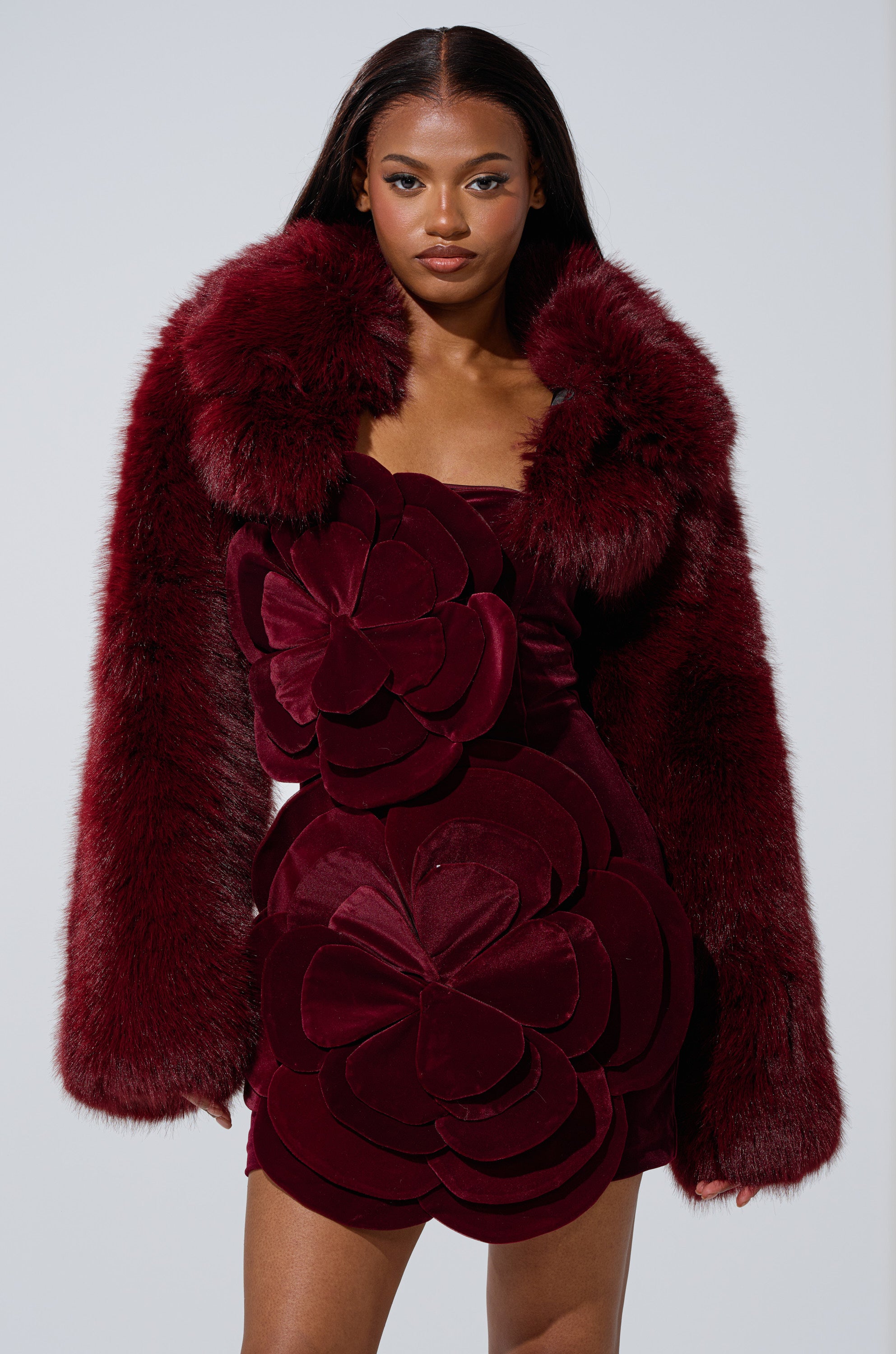 A woman poses confidently in the LEILA ULTRA CROP FAUX FUR IN BURGUNDY jacket, paired with a matching velvet dress featuring bold, layered floral designs, set against a plain light background.