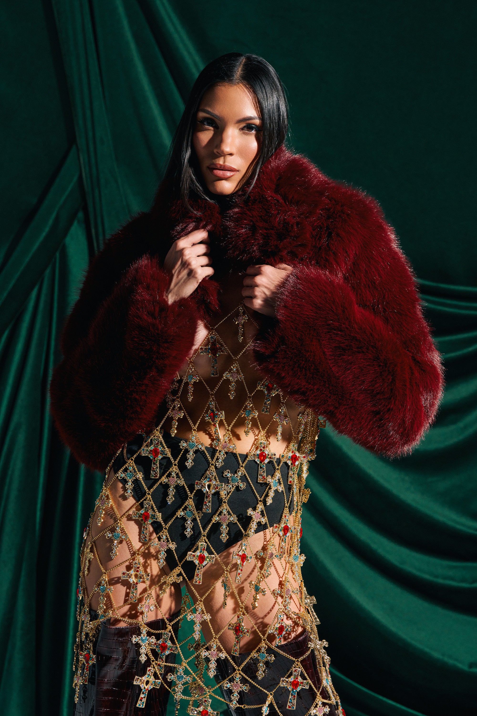 A woman stands before a dark green backdrop, confidently looking at the camera in the LEILA ULTRA CROP FAUX FUR IN BURGUNDY, paired with a sheer gold dress decorated with jewel-like crosses. She holds the jacket’s plush collar.