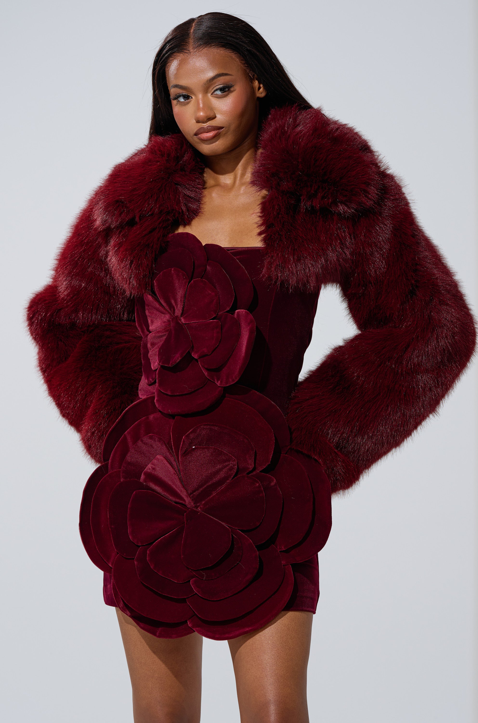 A woman confidently poses against a plain light background, wearing the AKIRA Label LEILA ULTRA CROP FAUX FUR IN BURGUNDY with oversized sleeves, paired with a matching mini dress decorated with bold, layered flower appliques.
