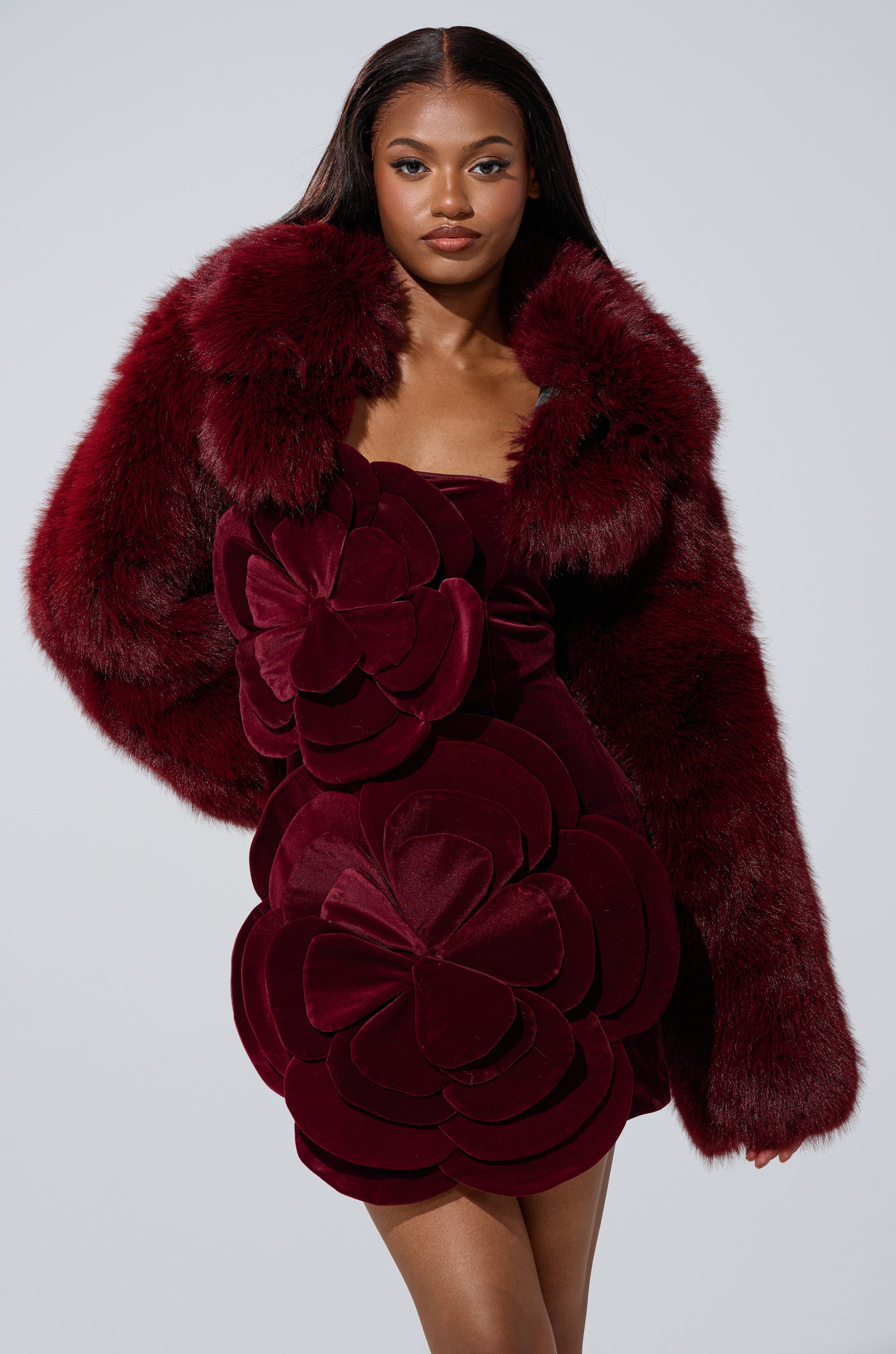 A woman poses confidently against a plain light background in the LEILA ULTRA CROP FAUX FUR IN BURGUNDY, styled with a matching short dress featuring large, layered flower-shaped fabric details.