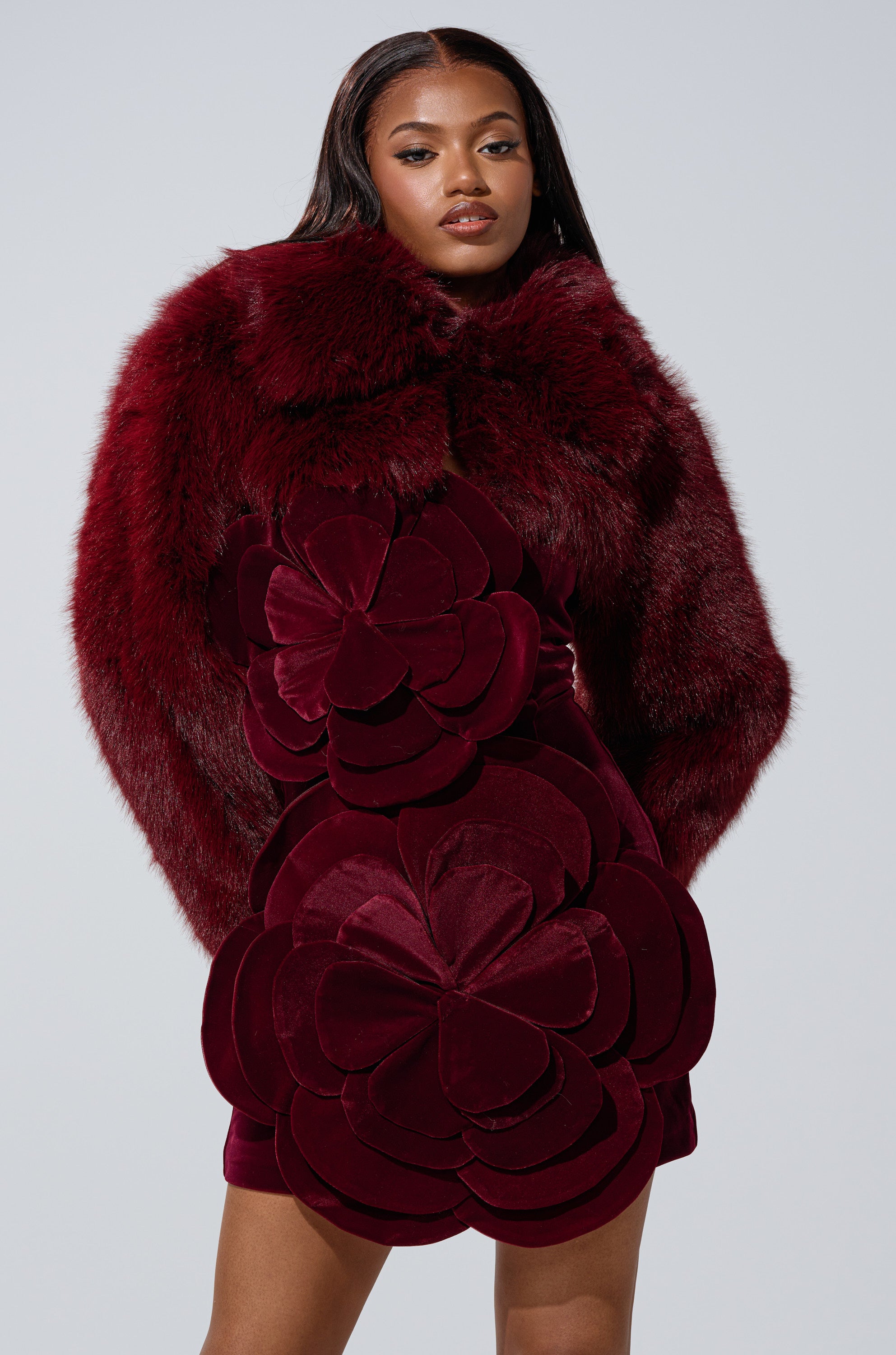 A woman poses confidently against a light gray background in the LEILA ULTRA CROP FAUX FUR IN BURGUNDY, styled with a deep burgundy dress featuring large, layered flower shapes for a bold, modern look.