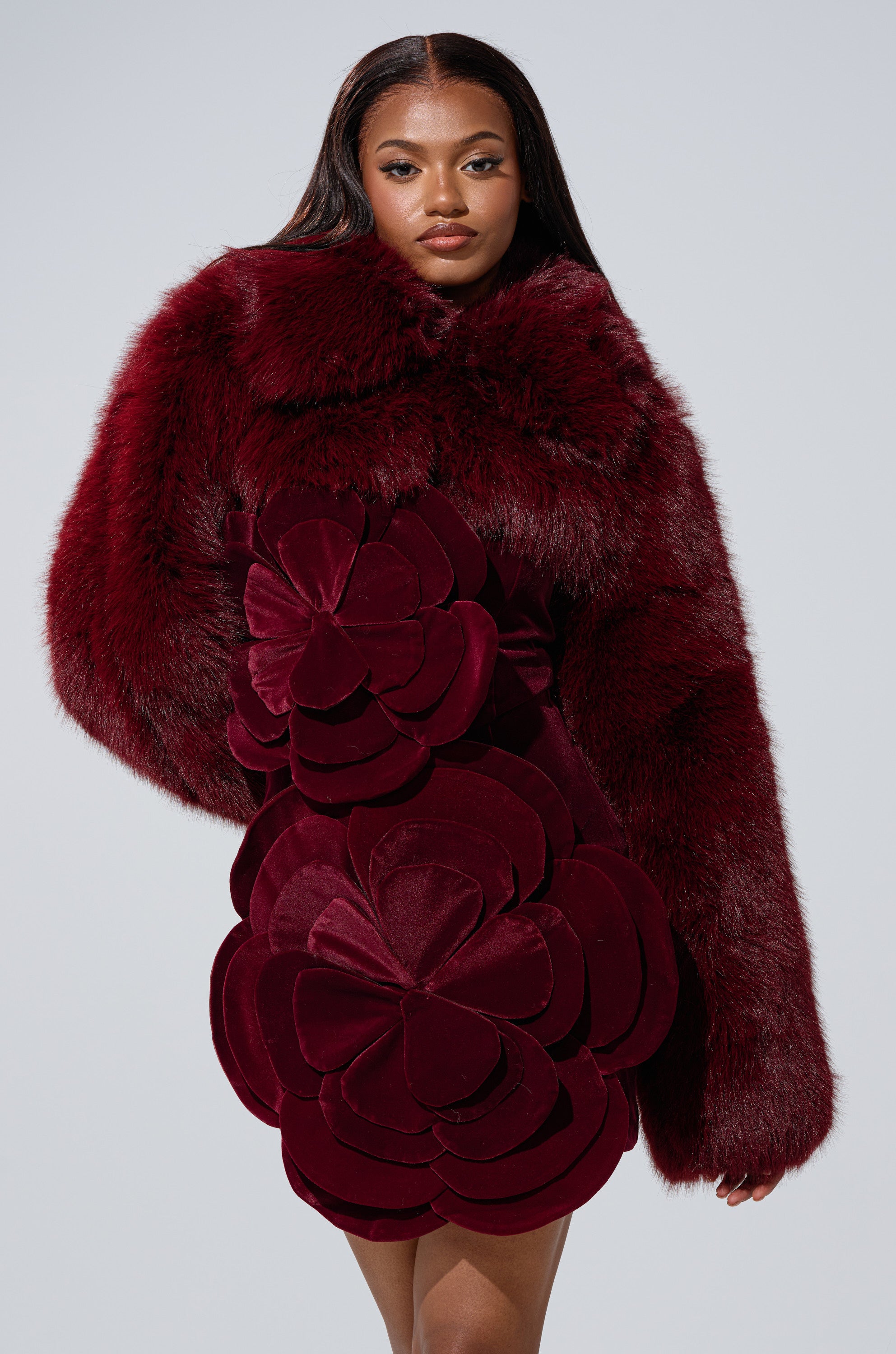 A woman stands confidently against a light gray background, wearing the LEILA ULTRA CROP FAUX FUR IN BURGUNDY—a luxurious jacket featuring oversized floral patterns and ultra-cropped faux fur sleeves.