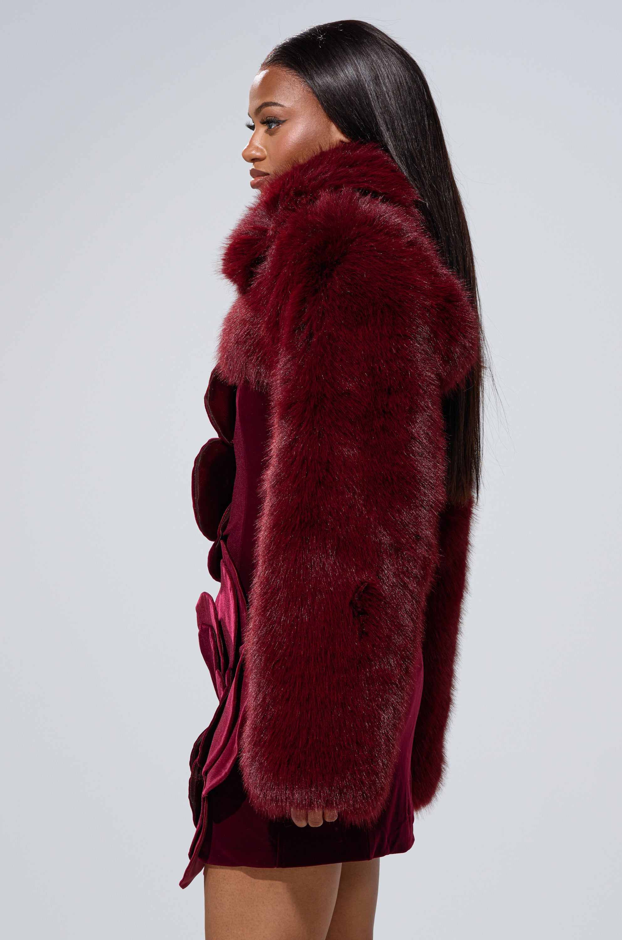 A woman with long straight hair wears the LEILA ULTRA CROP FAUX FUR IN BURGUNDY jacket and matching dress, standing in profile against a plain light gray background.