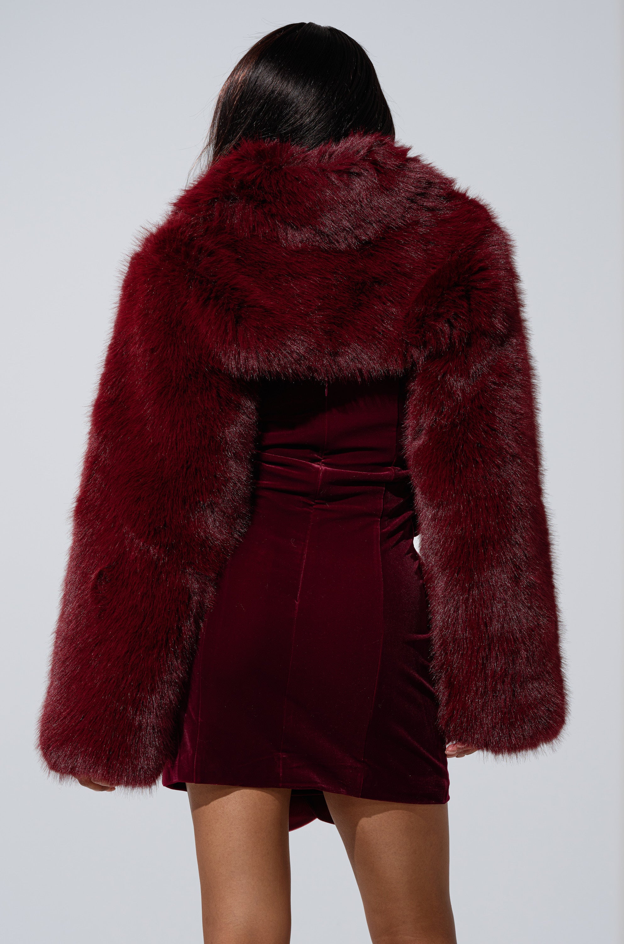 A person faces away, showcasing the LEILA ULTRA CROP FAUX FUR IN BURGUNDY jacket over a matching velvet dress. The photo displays their shoulders to thighs against a plain light background.