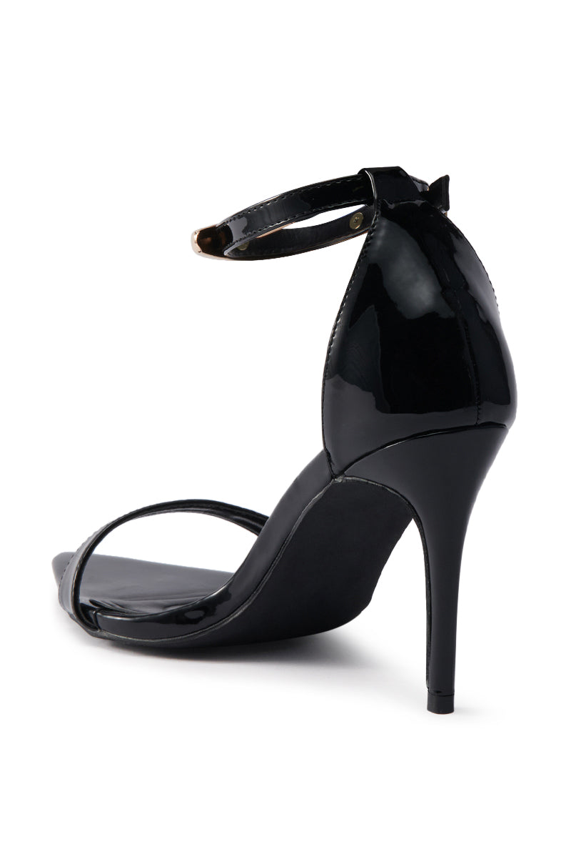 The AKIRA NEMTI BLACK STILETTO SANDAL, with a patent leather upper, adjustable ankle strap, and open toe, is displayed from a back-right angle against a white background.