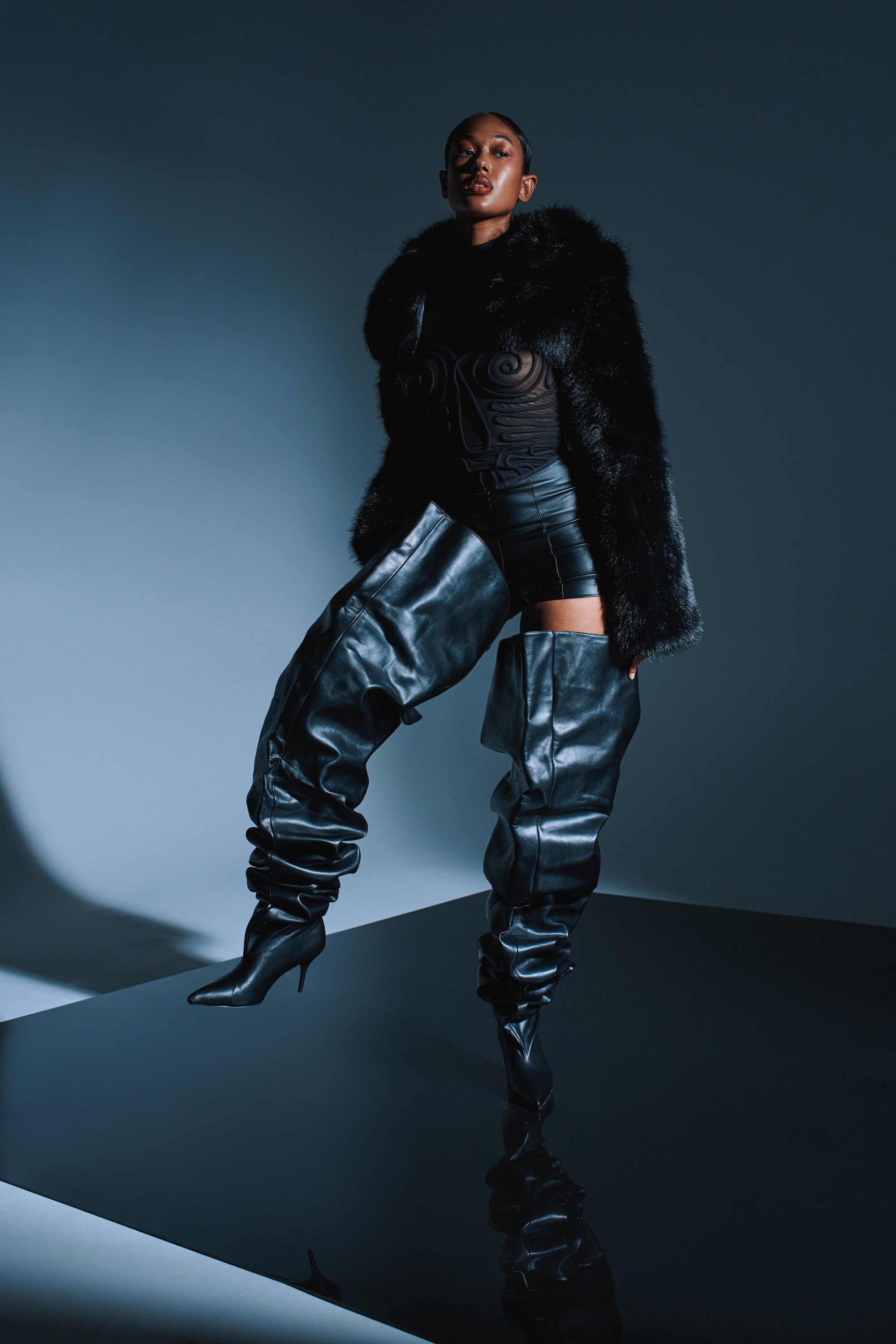 A person poses confidently in a black sheer top, faux fur jacket, and AZALEA WANG NEMY BLACK KITTEN HEEL THIGH HIGH BOOT, standing on a glossy surface against a dark background.