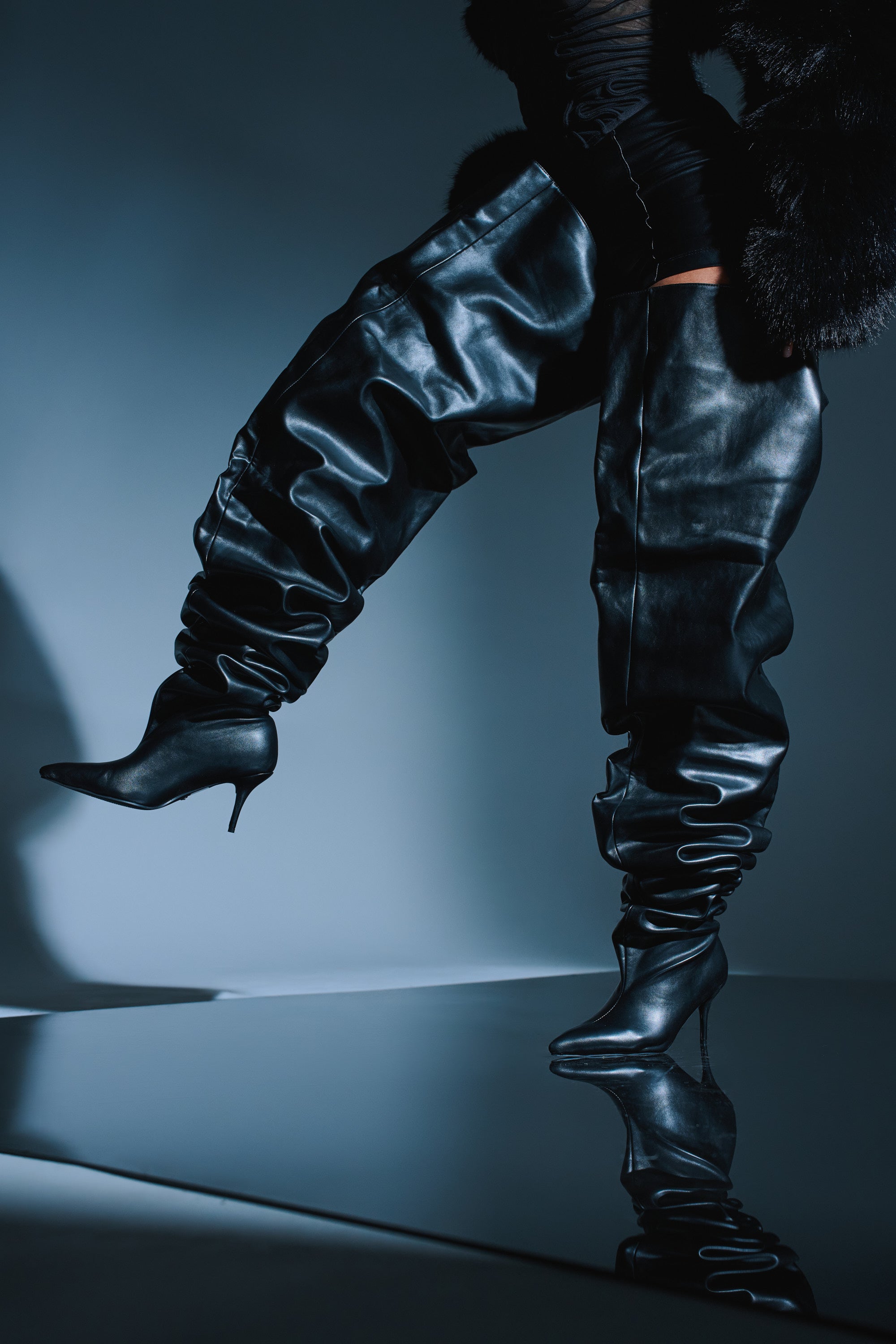 A person wearing the AZALEA WANG NEMY BLACK KITTEN HEEL THIGH HIGH BOOT and a black fur coat poses with one leg lifted, creating dramatic shadows on a glossy, reflective surface.