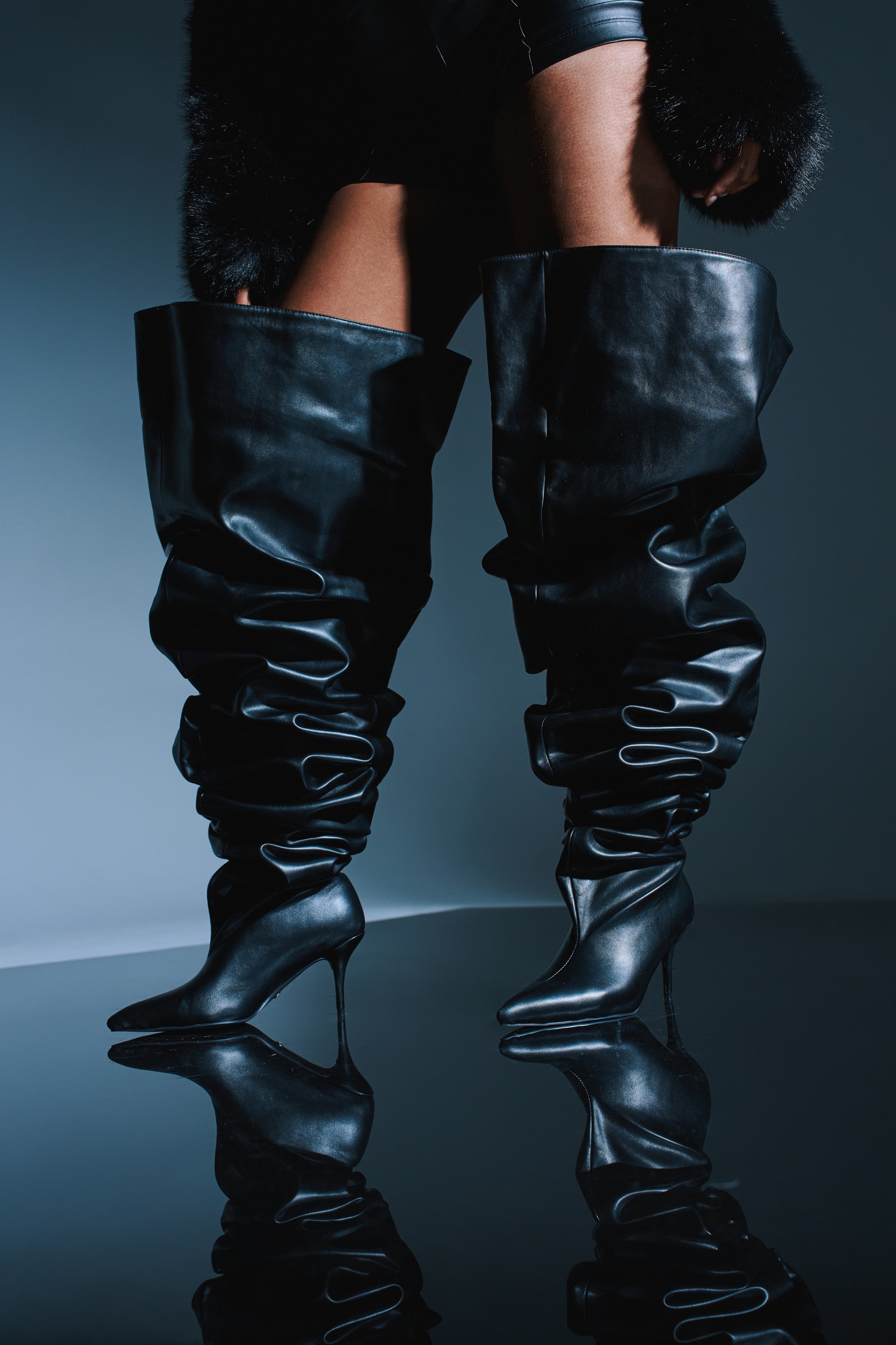 A person wearing AZALEA WANG NEMY Black Kitten Heel Thigh High Boots, black shorts, and a fur coat stands on a reflective surface against a dark background.