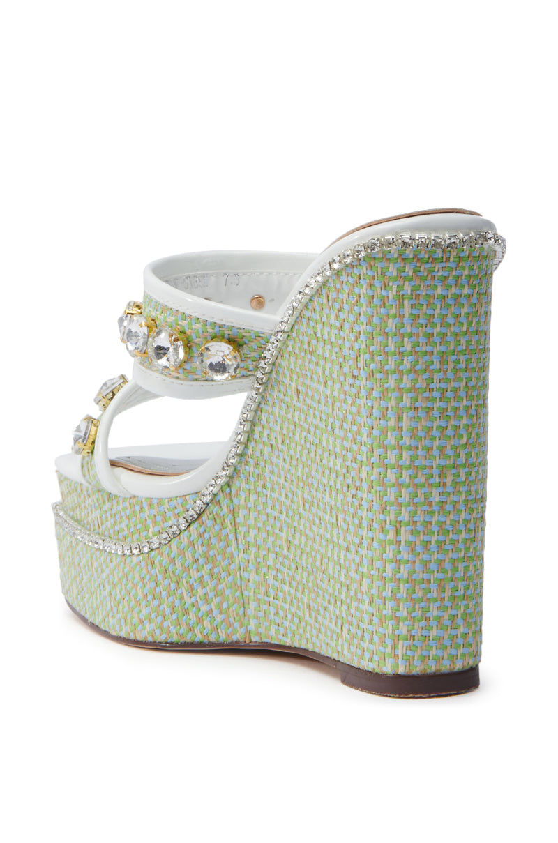 The image displays the AZALEA WANG NIOMA GREEN EMBELLISHED WEDGE SANDAL, featuring a chunky wedge heel, woven green and blue textures, crystal-adorned straps, and decorative trim on the heel edge.
