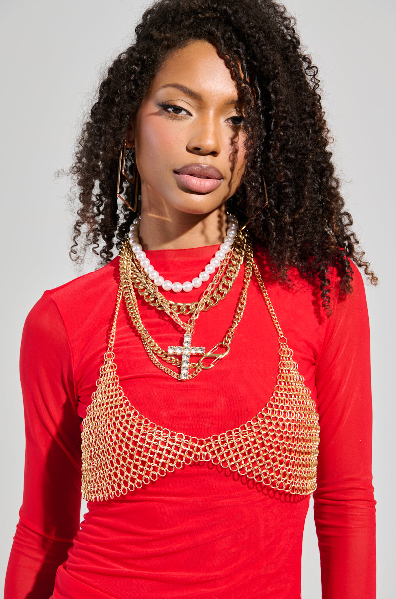 A person with curly hair wears a vibrant red top adorned with multiple layered necklaces, such as gold chains and pearls. Among the accessories is the CLASSIC BODY CHAIN, characterized by its gold chain link featuring a cross pendant. The background is softly muted in a neutral, light color.