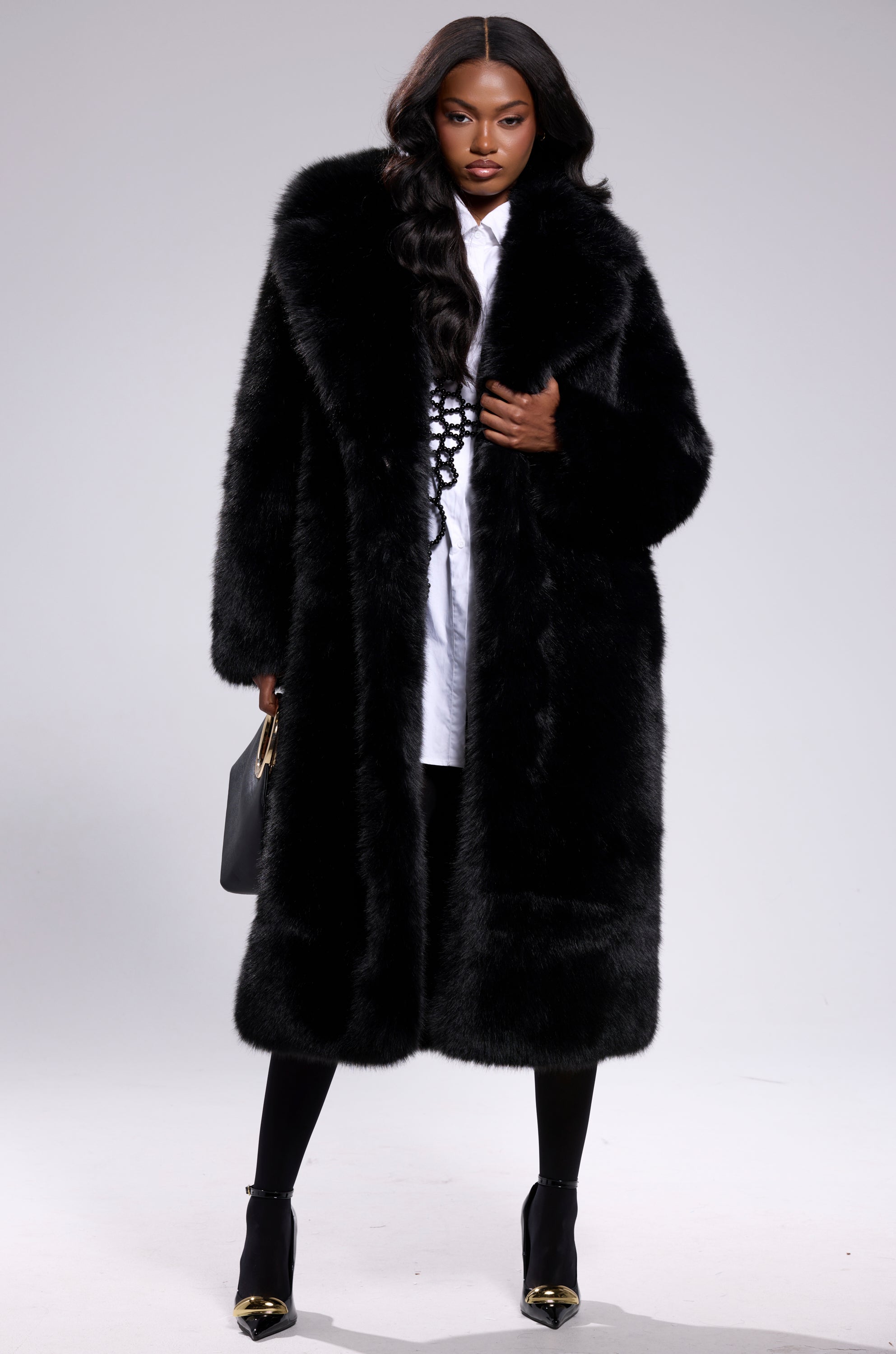 A person wearing the OSLO FAUX FUR COAT IN BLACK over a white shirt, black leggings, and gold-tipped black heels, holding a black handbag, stands against a plain light gray background.