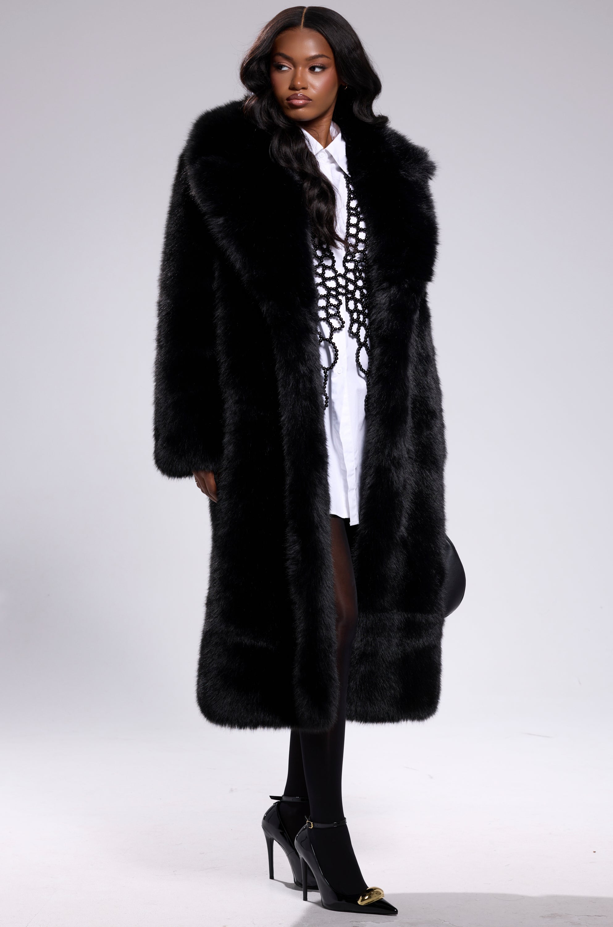 A woman wears the OSLO FAUX FUR COAT IN BLACK over a white shirt dress, black knit corset, black tights, and black heels, standing against a plain light background.