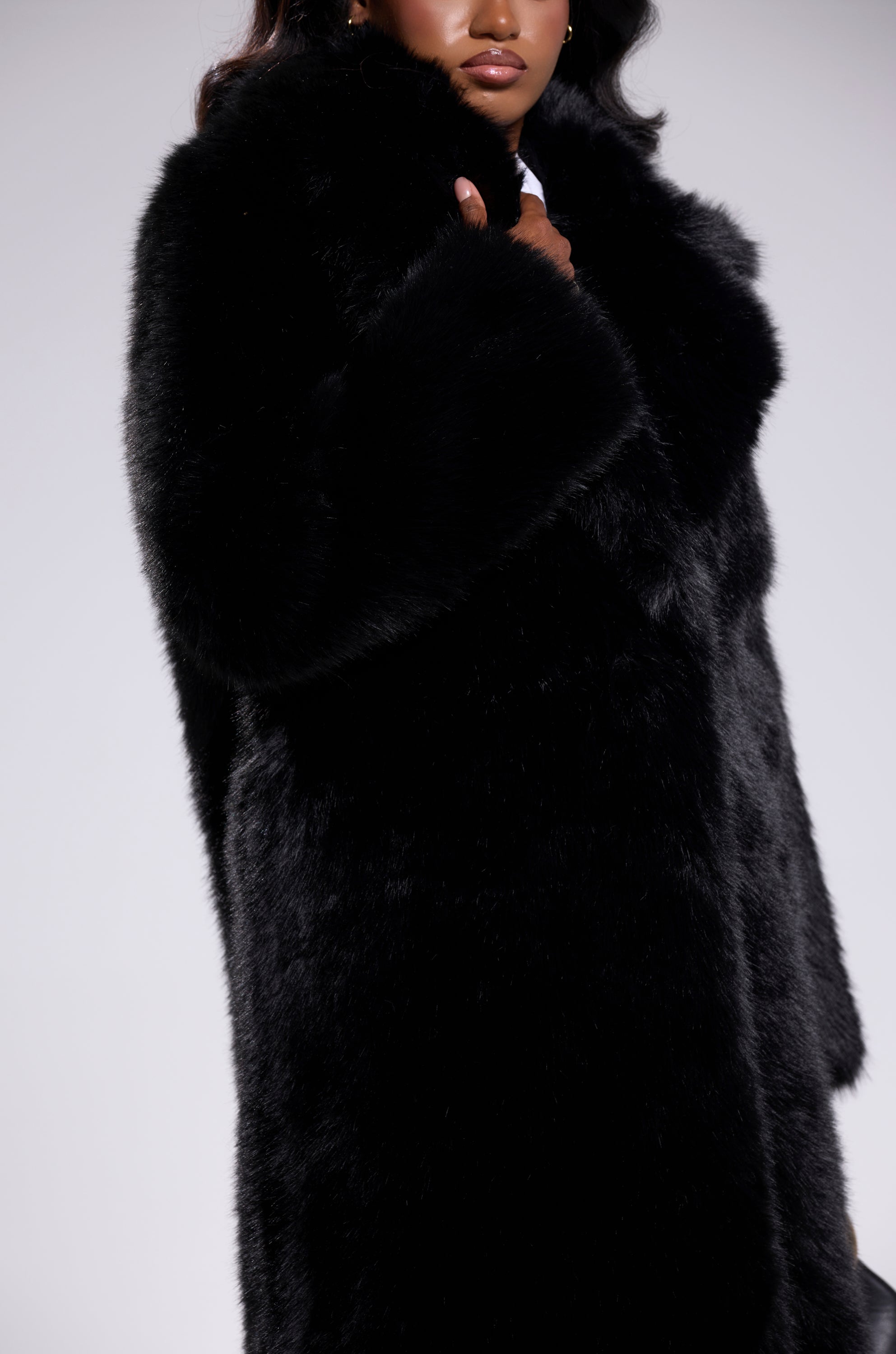 A person models the OSLO FAUX FUR COAT IN BLACK—a luxurious, long black faux fur coat with an upturned collar and arms partially crossed—set against a light gray background with their face partially visible.