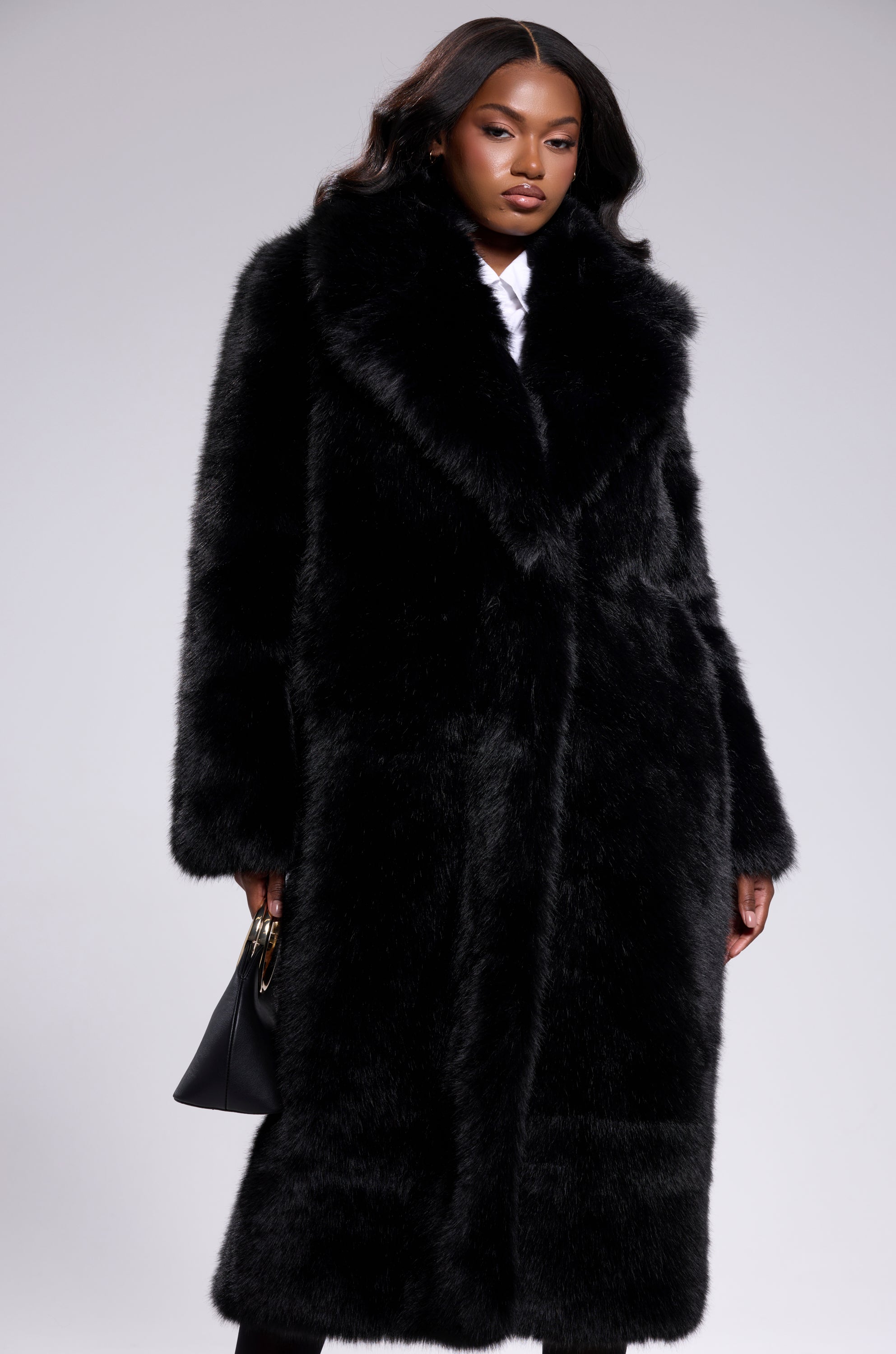 Wearing the OSLO FAUX FUR COAT IN BLACK, a woman with long, wavy hair and a confident look holds a black handbag against a plain, light gray background.
