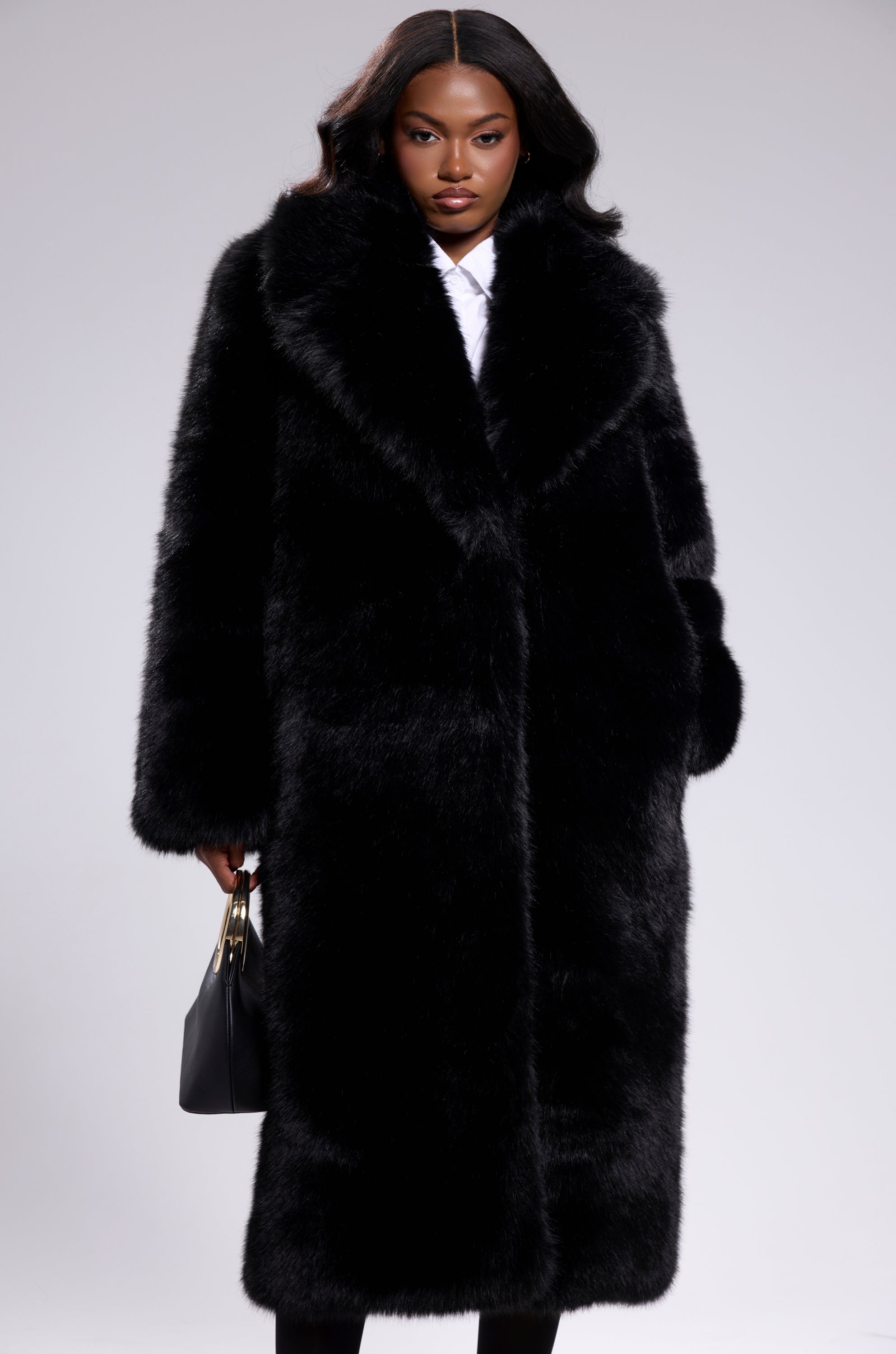 A woman with long, wavy hair faces forward in a plain light gray setting, wearing the OSLO FAUX FUR COAT IN BLACK over a white shirt and holding a black handbag.