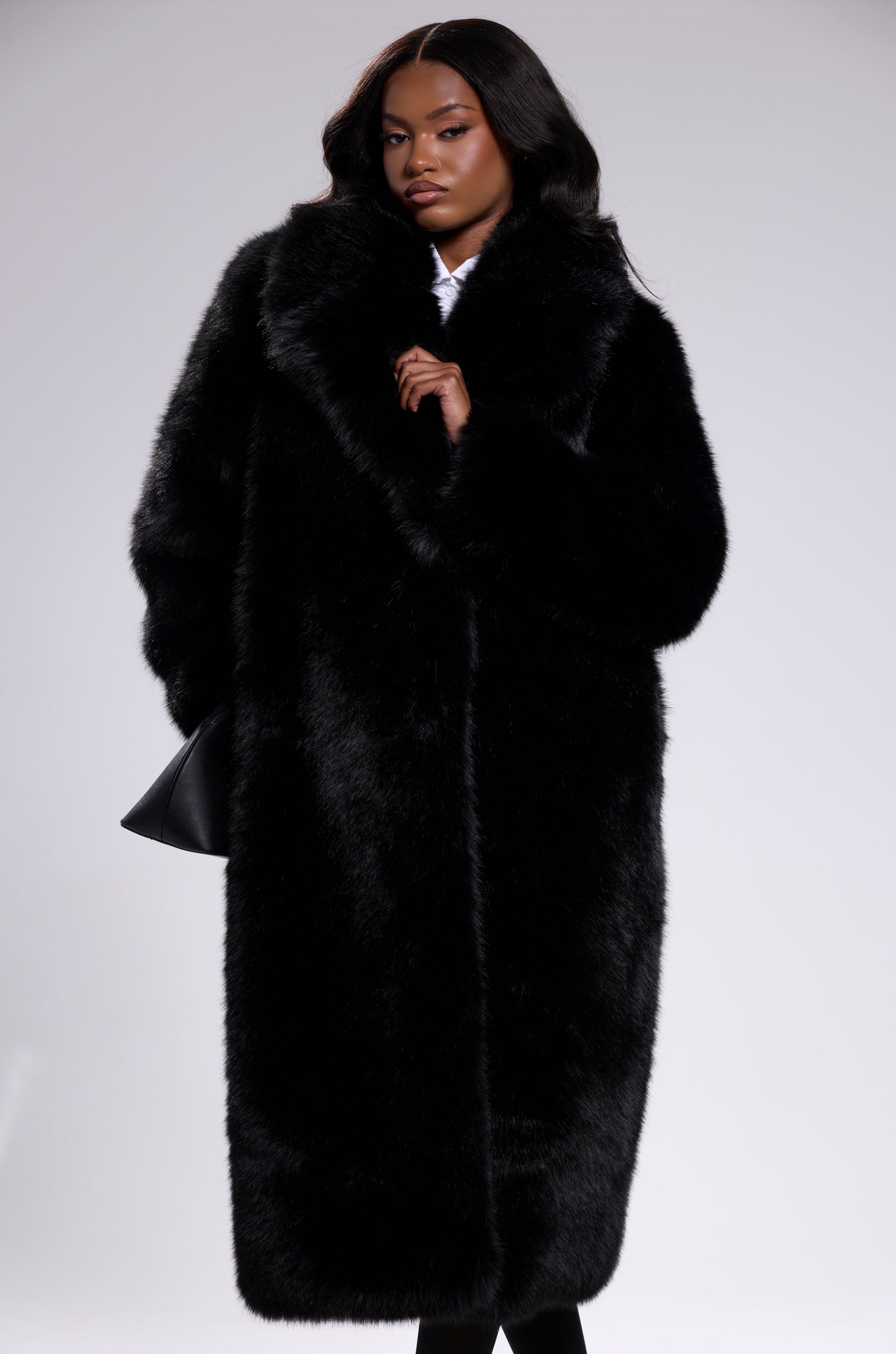 A woman stands against a light grey background wearing the OSLO FAUX FUR COAT IN BLACK, holding the oversized coat closed with one hand while her other hand rests at her side.