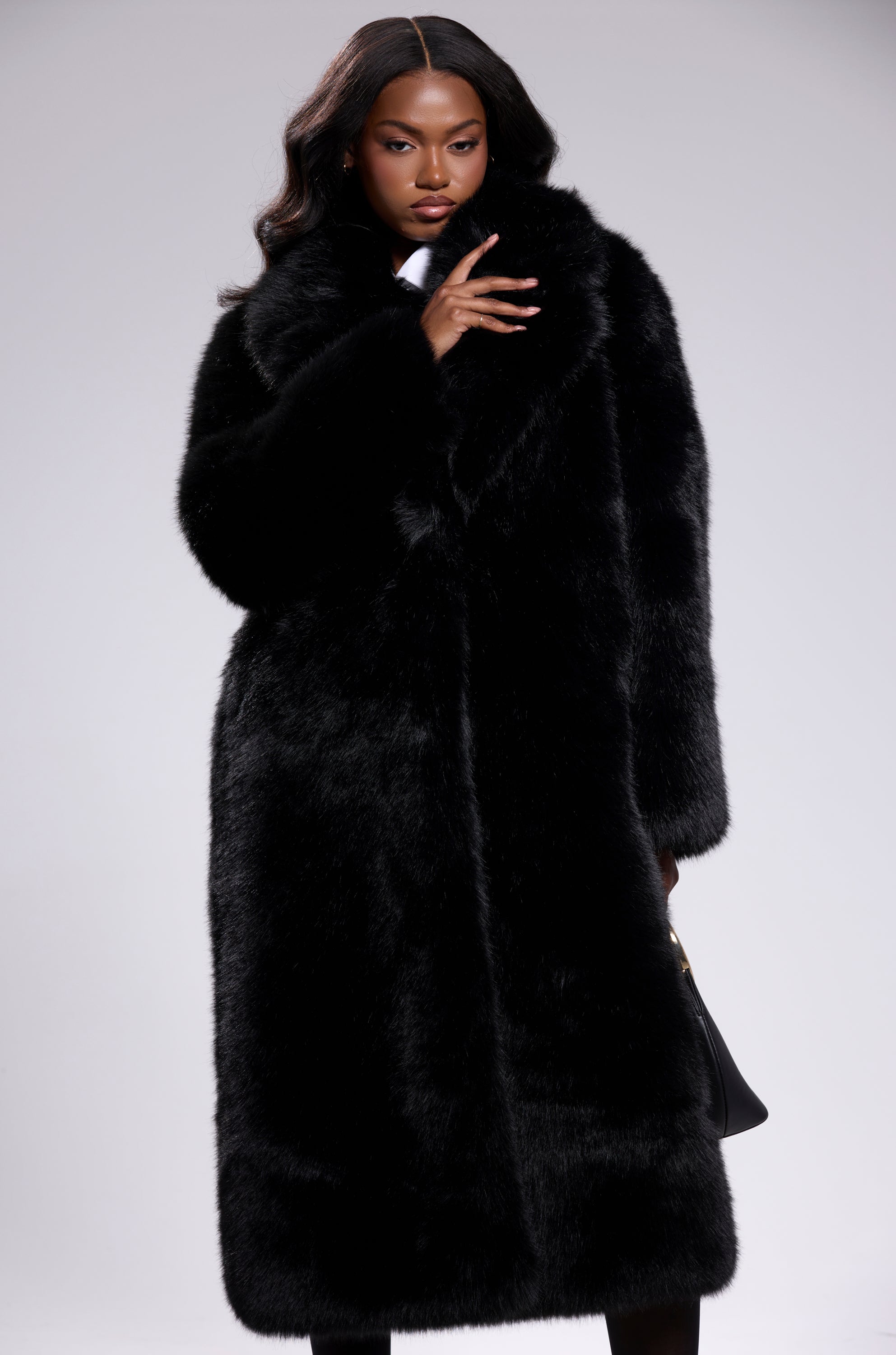 A woman with long, wavy hair poses confidently in the OSLO FAUX FUR COAT IN BLACK, standing against a light gray background. She holds the coat near her face and carries a black handbag by her side.
