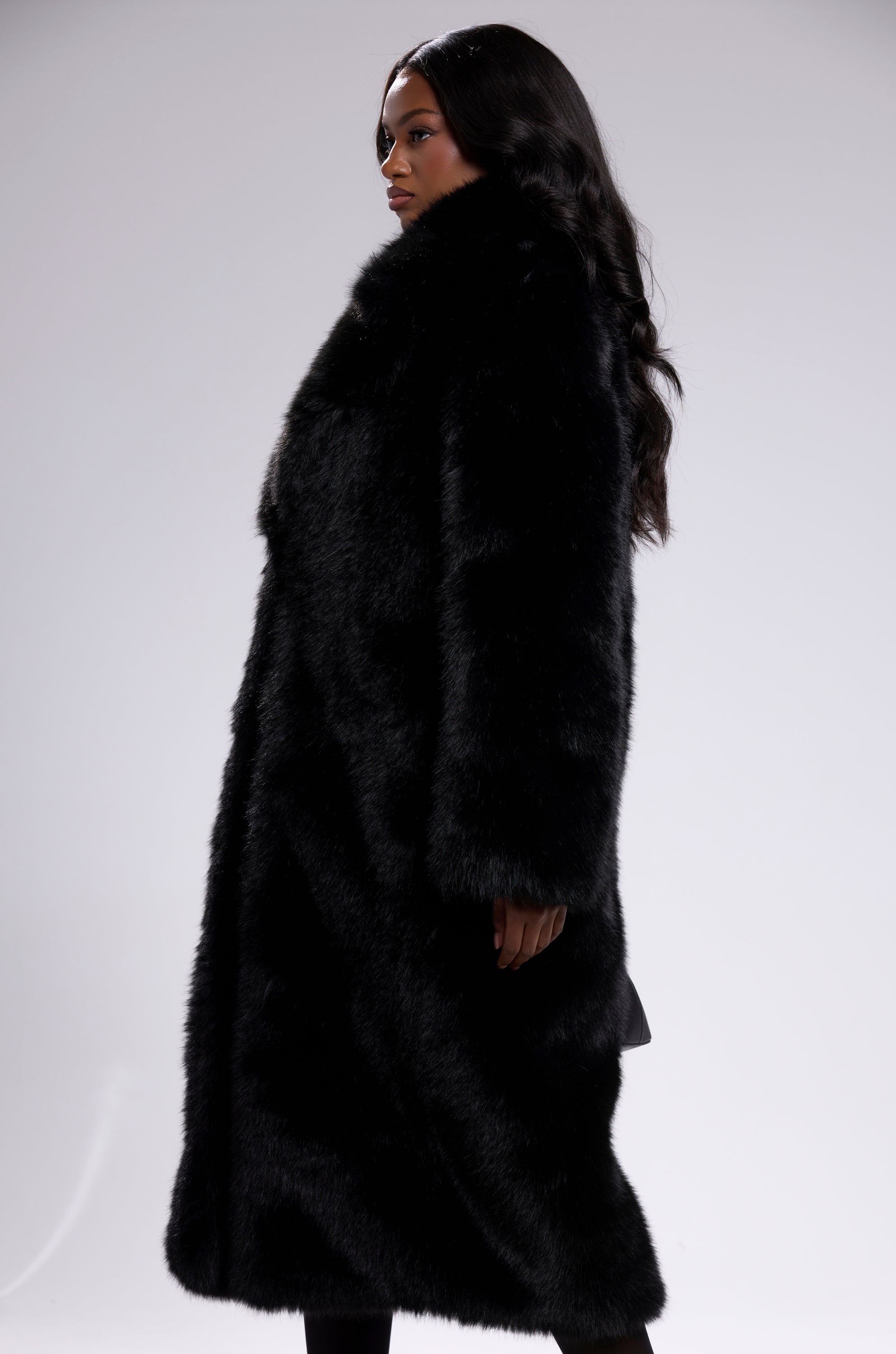 A woman with long, wavy hair stands in profile, modeling the OSLO FAUX FUR COAT IN BLACK against a plain gray background.