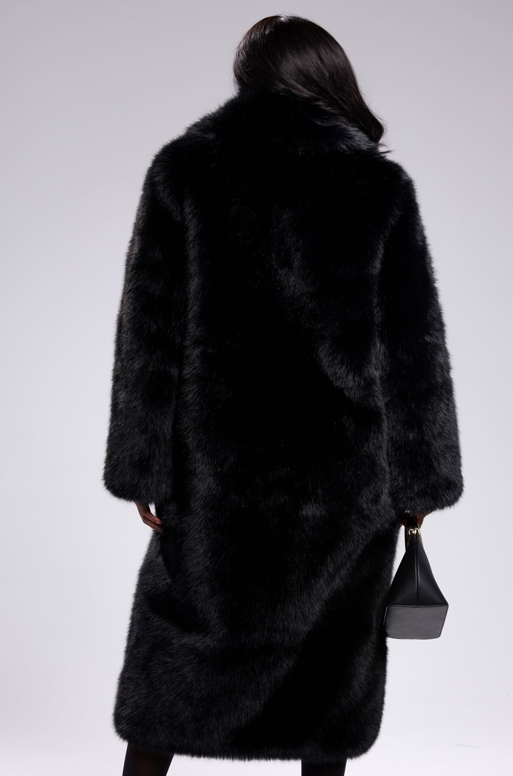 A person with long dark hair stands with their back to the camera, wearing the OSLO FAUX FUR COAT IN BLACK and holding a small black handbag against a light gray background.
