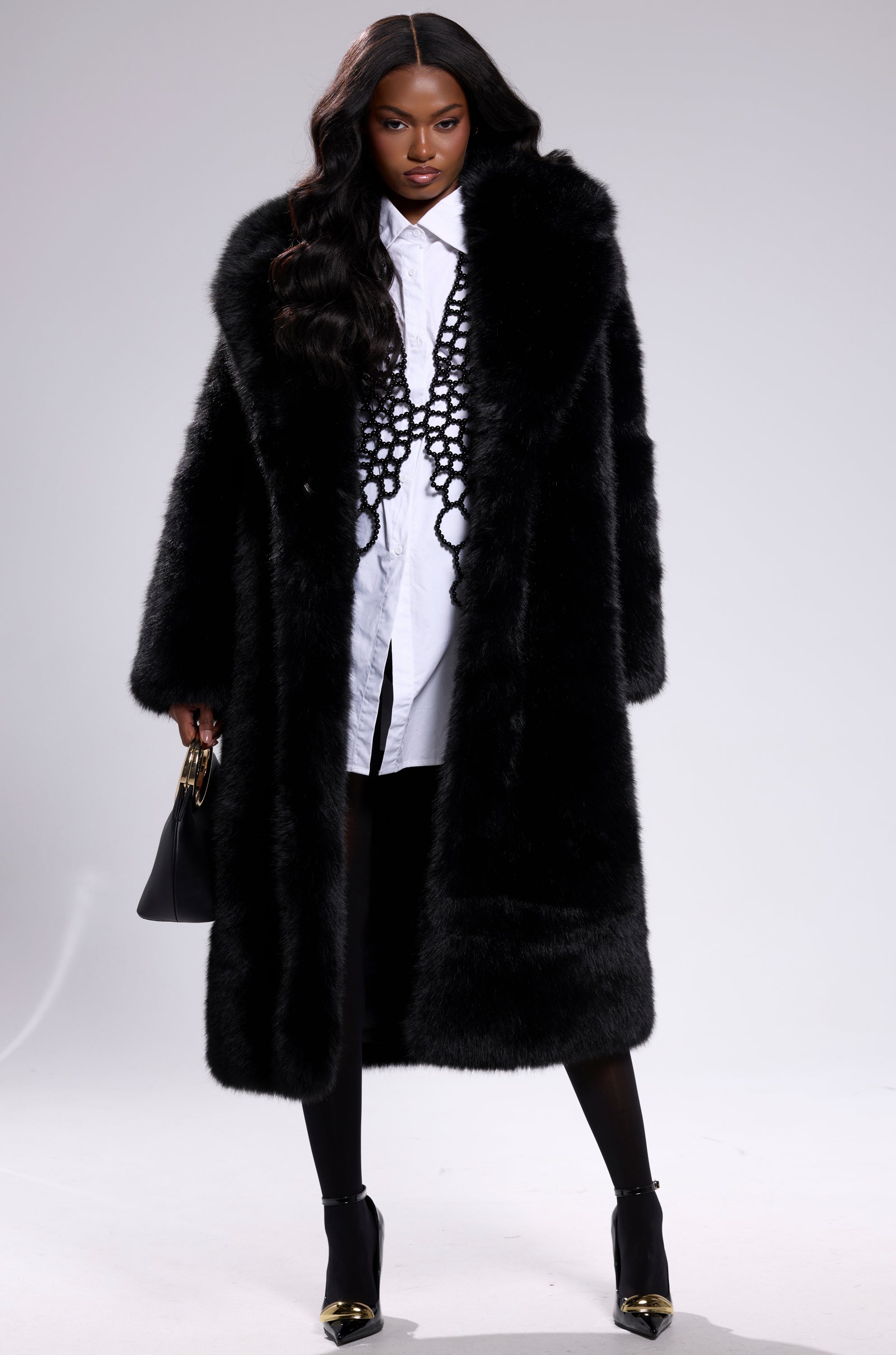 A woman stands confidently in the OSLO FAUX FUR COAT IN BLACK over a white shirt, black skirt, tights, ankle boots, and a bold necklace. She carries a black handbag and her long wavy hair completes the chic look.