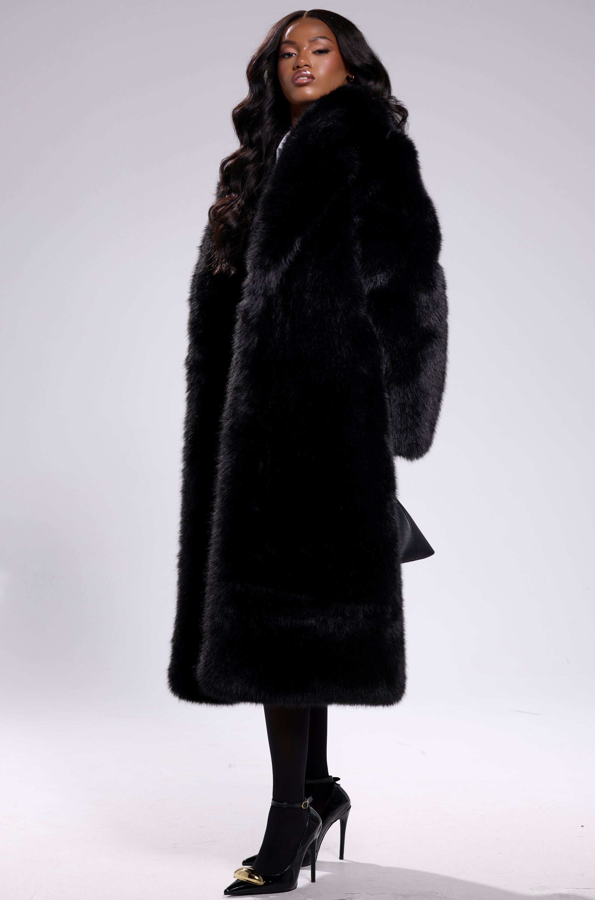 A person with long dark hair models the OSLO FAUX FUR COAT IN BLACK, paired with black tights and gold-accented heels, standing against a plain light gray background.