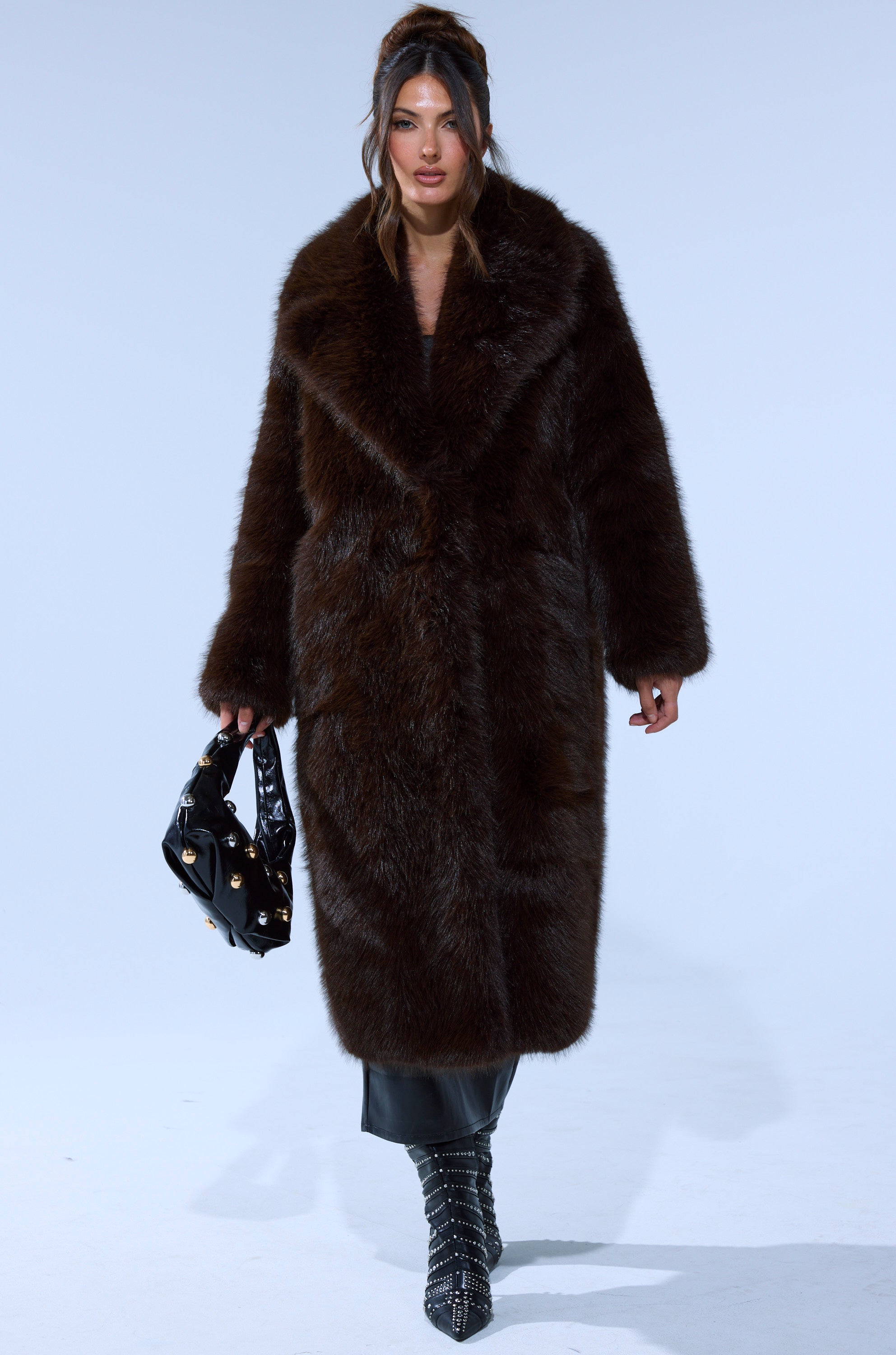 A woman in the OSLO FAUX FUR COAT IN CHOCOLATE, a black skirt, studded black boots, and holding a black handbag with gold accents walks forward against a plain light background.