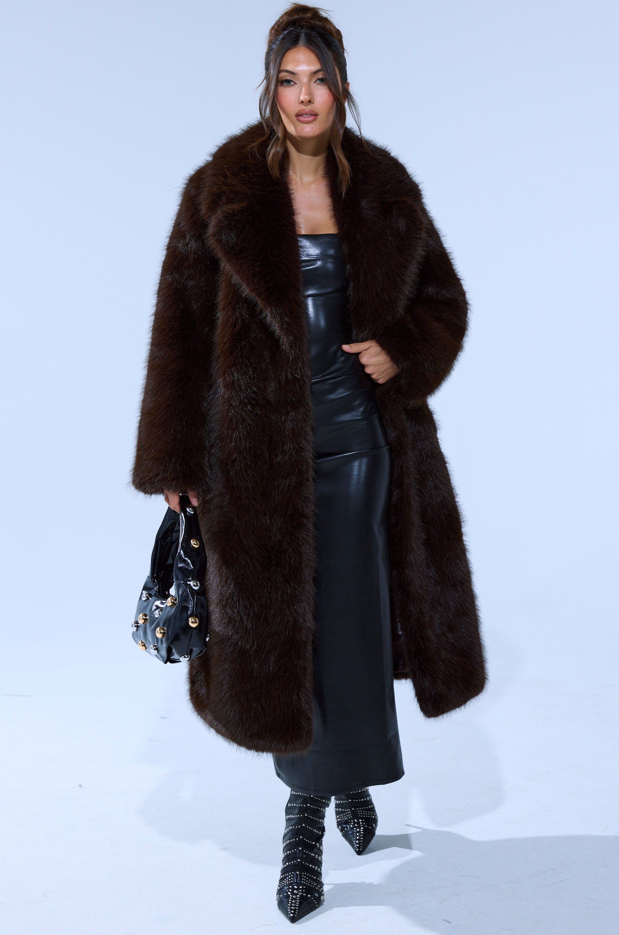 A woman wears the OSLO FAUX FUR COAT IN CHOCOLATE over a black leather dress, holding a black handbag with gold accents and wearing pointed black studded boots, standing against a light background.