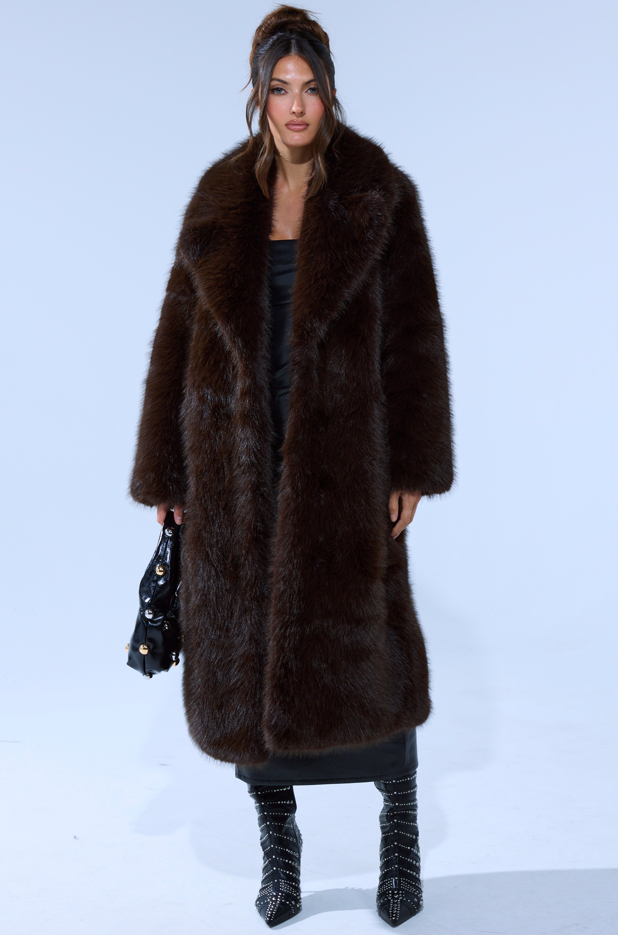 A woman stands against a plain background wearing the OSLO FAUX FUR COAT IN CHOCOLATE over a black outfit. She holds a black handbag, wears pointed studded boots, and her hair is styled half-up with loose waves.