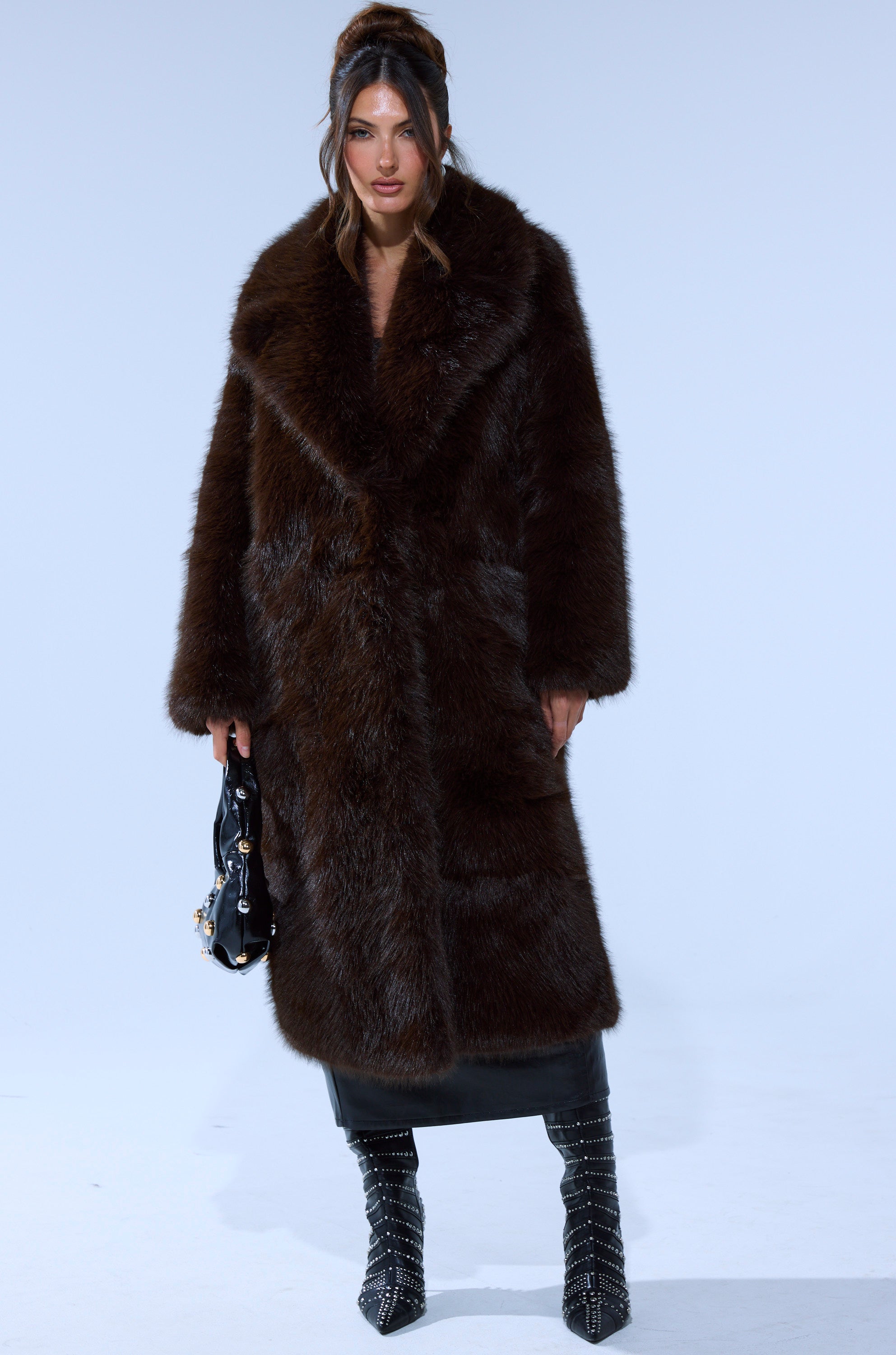 A woman stands against a plain background wearing the OSLO FAUX FUR COAT IN CHOCOLATE, paired with a black leather skirt, studded black boots, and a small black handbag. Her hair is styled in loose waves with some pulled back.