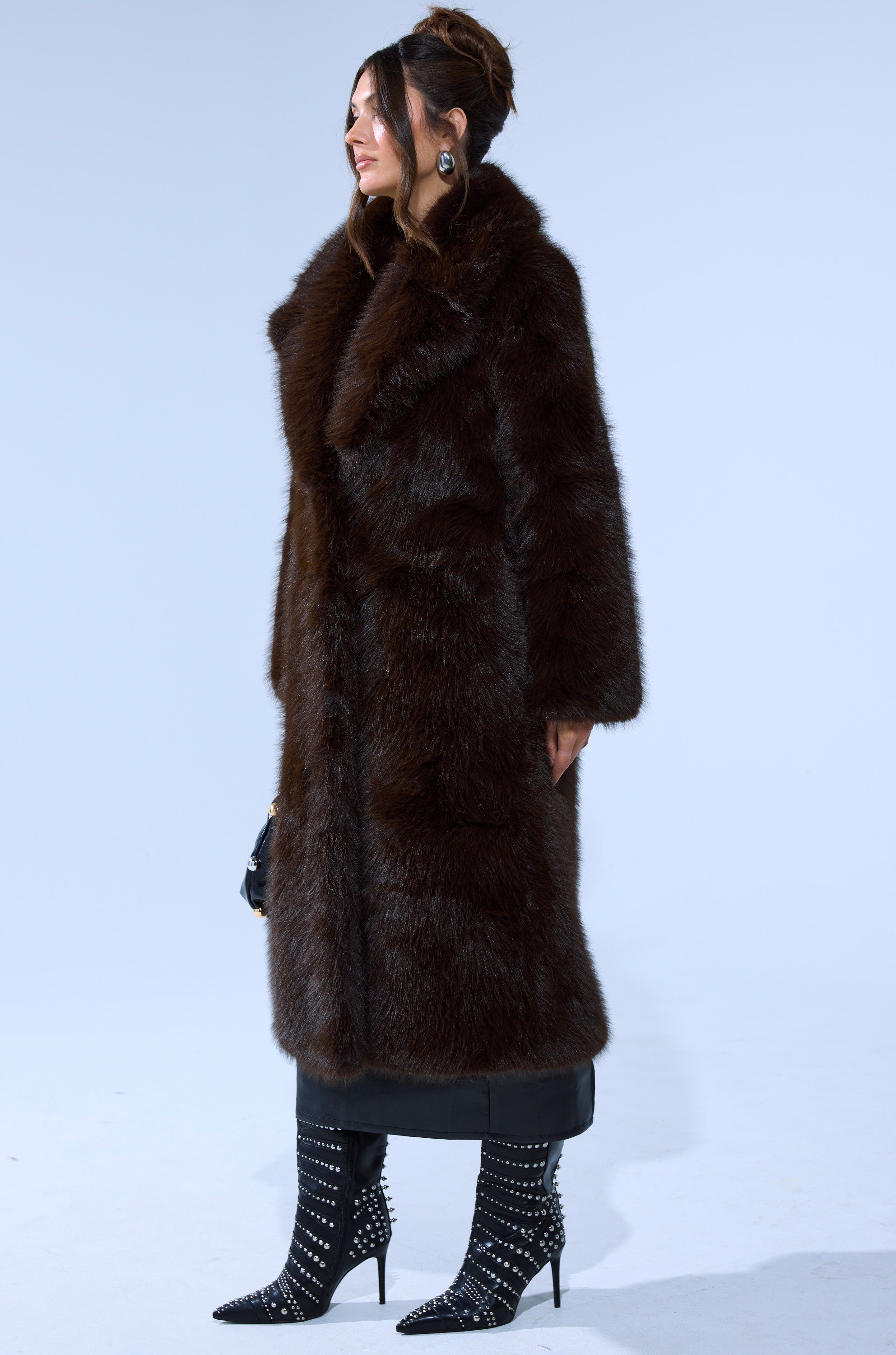 A woman in profile wears the OSLO FAUX FUR COAT IN CHOCOLATE with black studded high-heeled boots, black pants, and a small black bag, standing against a plain light background.