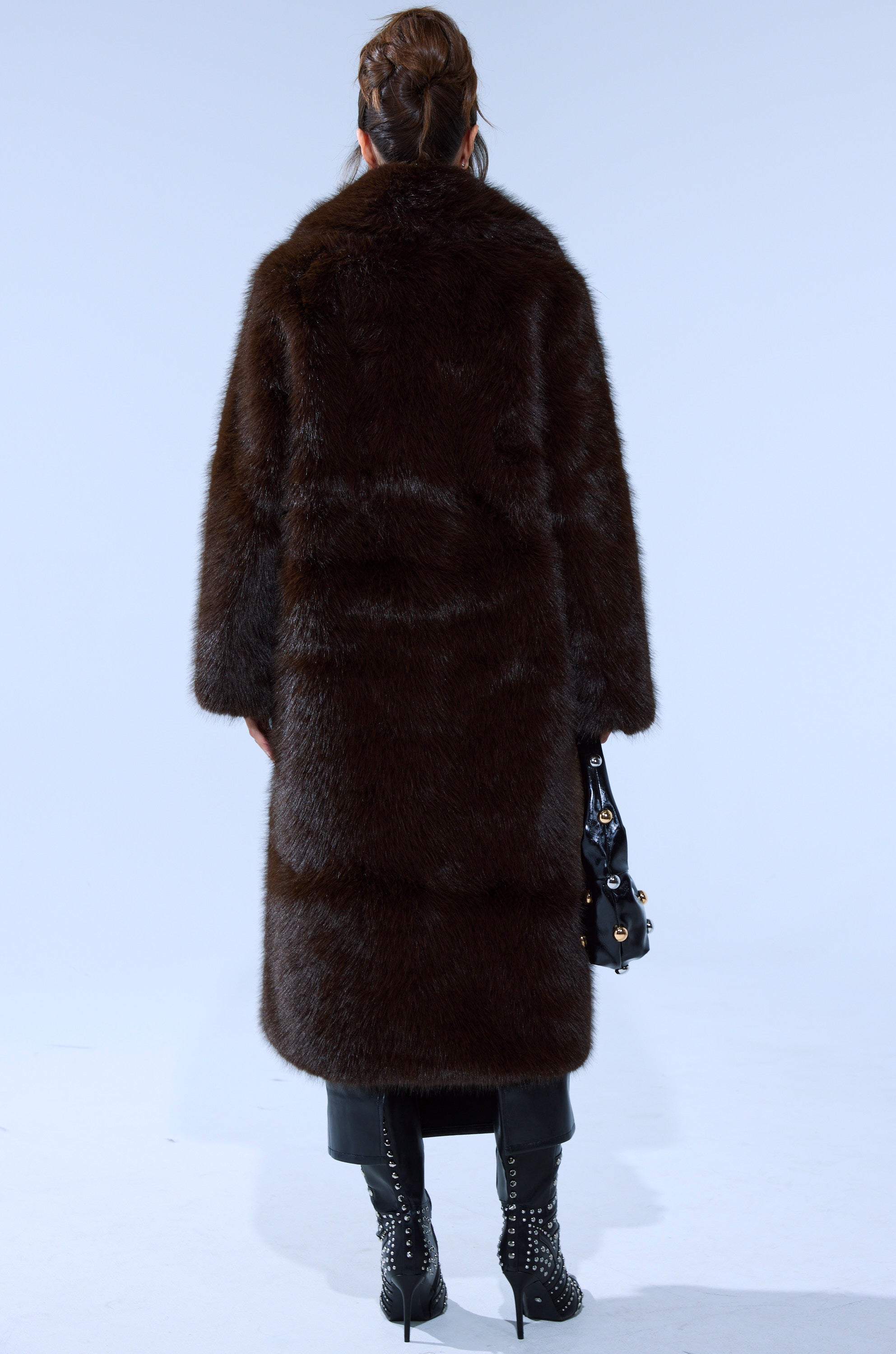 A person stands facing away in the OSLO FAUX FUR COAT IN CHOCOLATE, styled with black studded boots, black pants, a black handbag, and an updo hairstyle against a plain, light background.