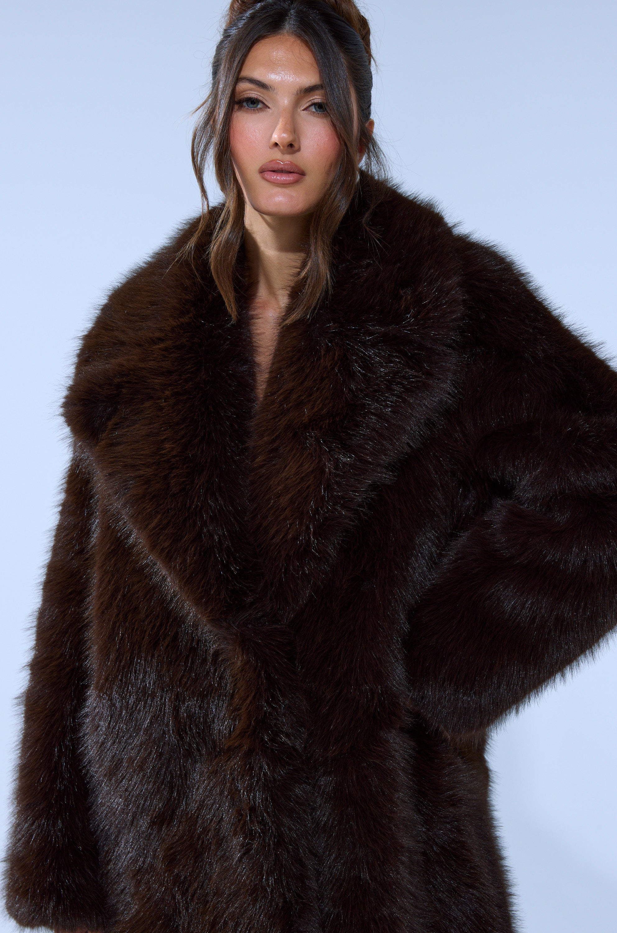 A woman with long brown hair wears the OSLO FAUX FUR COAT IN CHOCOLATE, standing confidently against a plain light blue background and gazing at the camera.