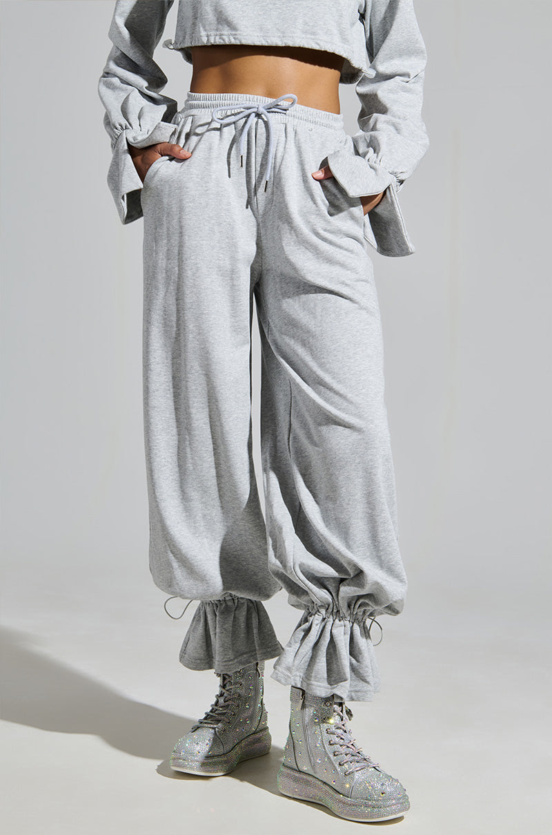 A person in a gray long-sleeved cropped top and matching drawstring sweatpants with ruffled ankle cuffs sports the AZALEA WANG OTIS RHINESTONE HIGH TOP SNEAKER IN SILVER, which sparkles against the plain gray background.