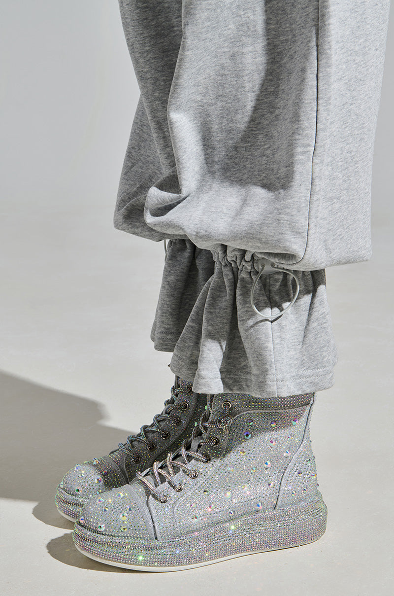 A person wearing gray pants with drawstring cuffs and the AZALEA WANG OTIS Rhinestone High Top Sneaker in Silver stands against a plain, minimalistic background.